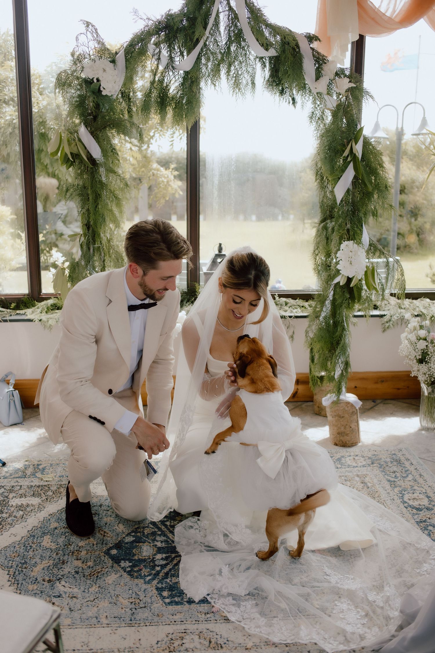A couple in wedding attire plays with a small dog during an intimate indoor photo session captured by a cotswolds wedding photographer.