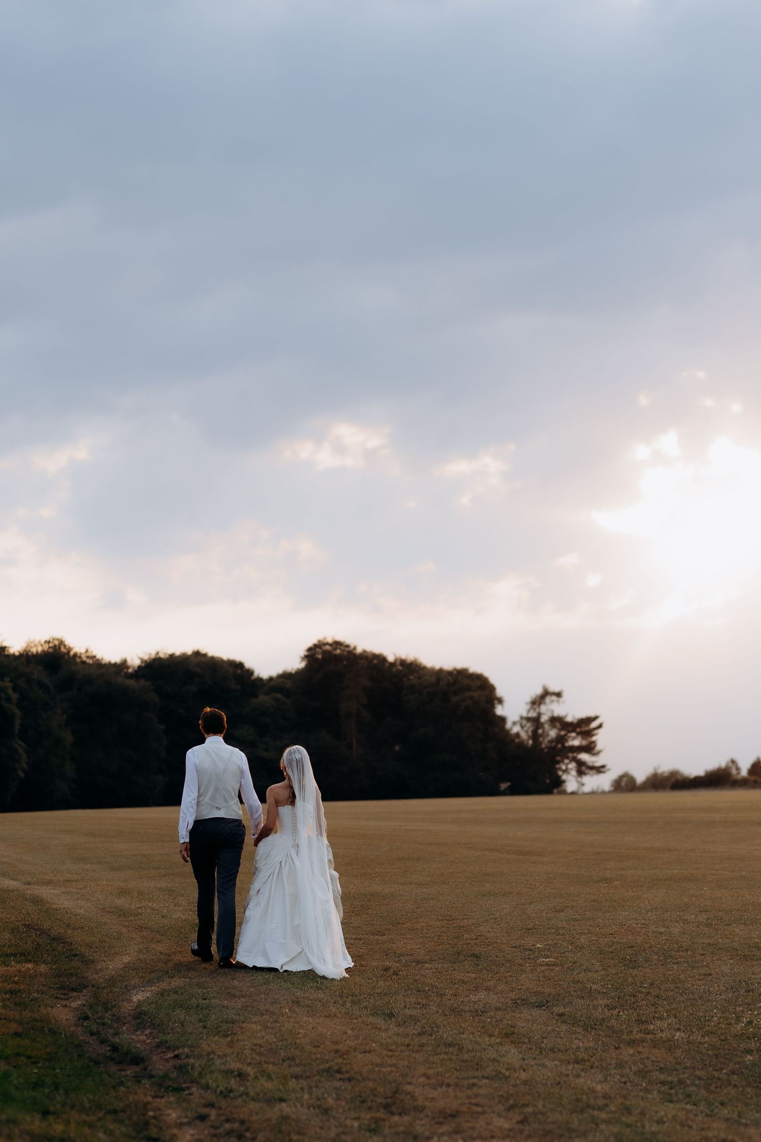 Bride and groom walk hand in hand across a golden field in the cotswolds under dramatic cloudy skies at sunset.