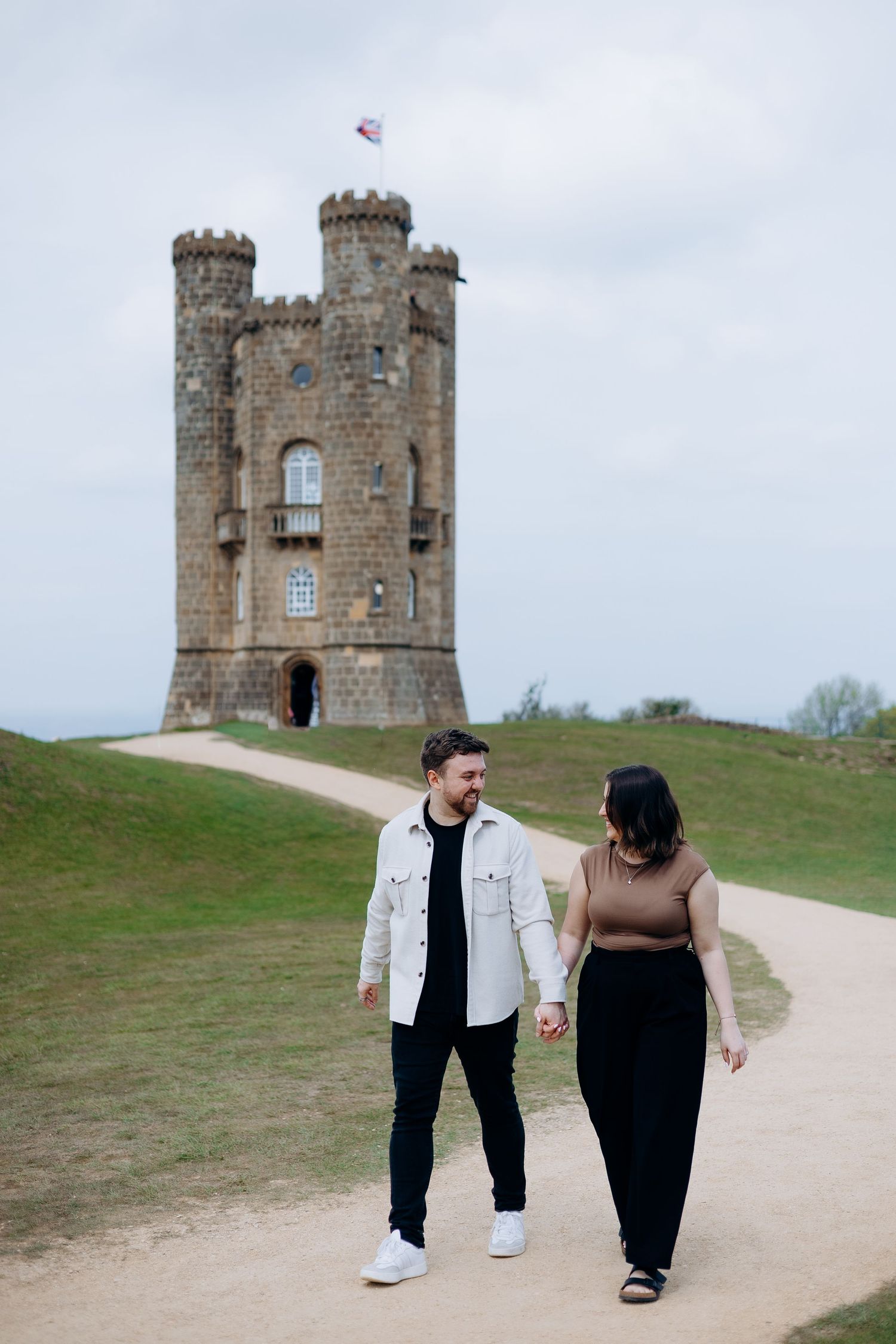 Couple holding hands walking on a path on their engagement photoshoot at Broadway Tower castle in the Cotswolds, England.