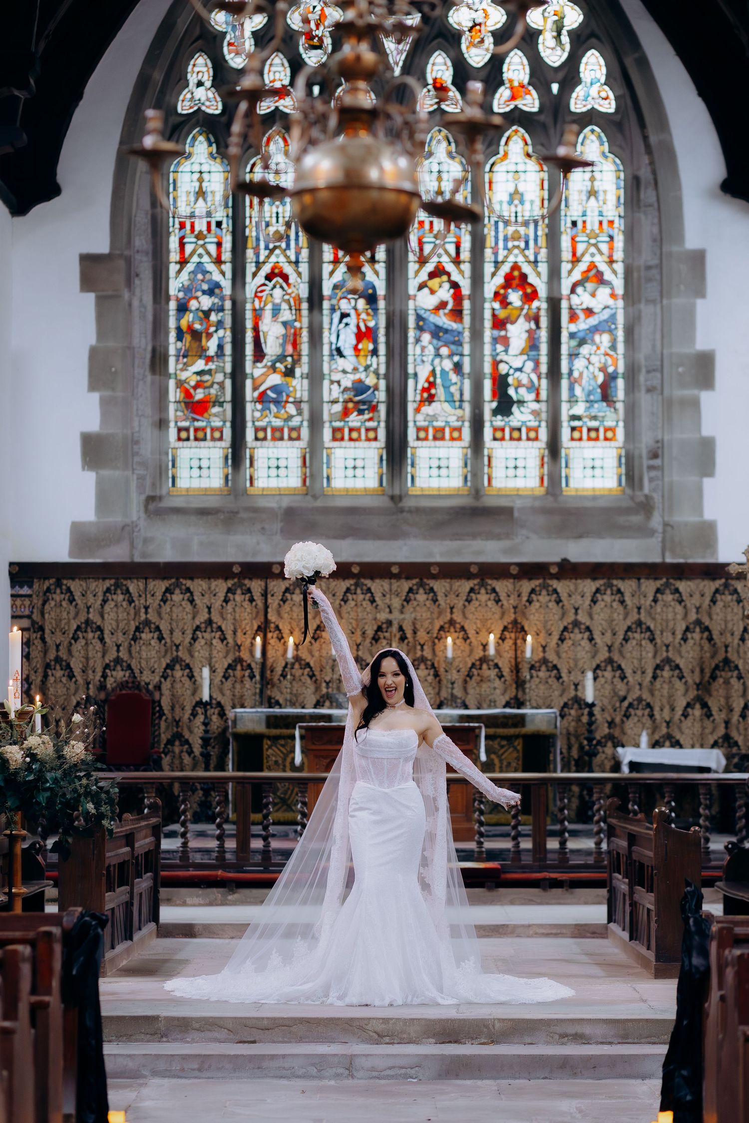 A bride in a white wedding dress kneels at the altar of a historic church with stained glass windows in the Cotswolds.