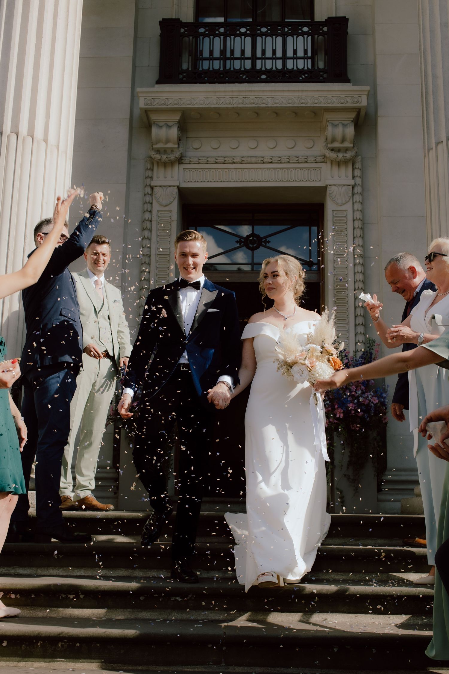 Wedding couple descending grand stone steps while guests throw confetti outside a London wedding venue.