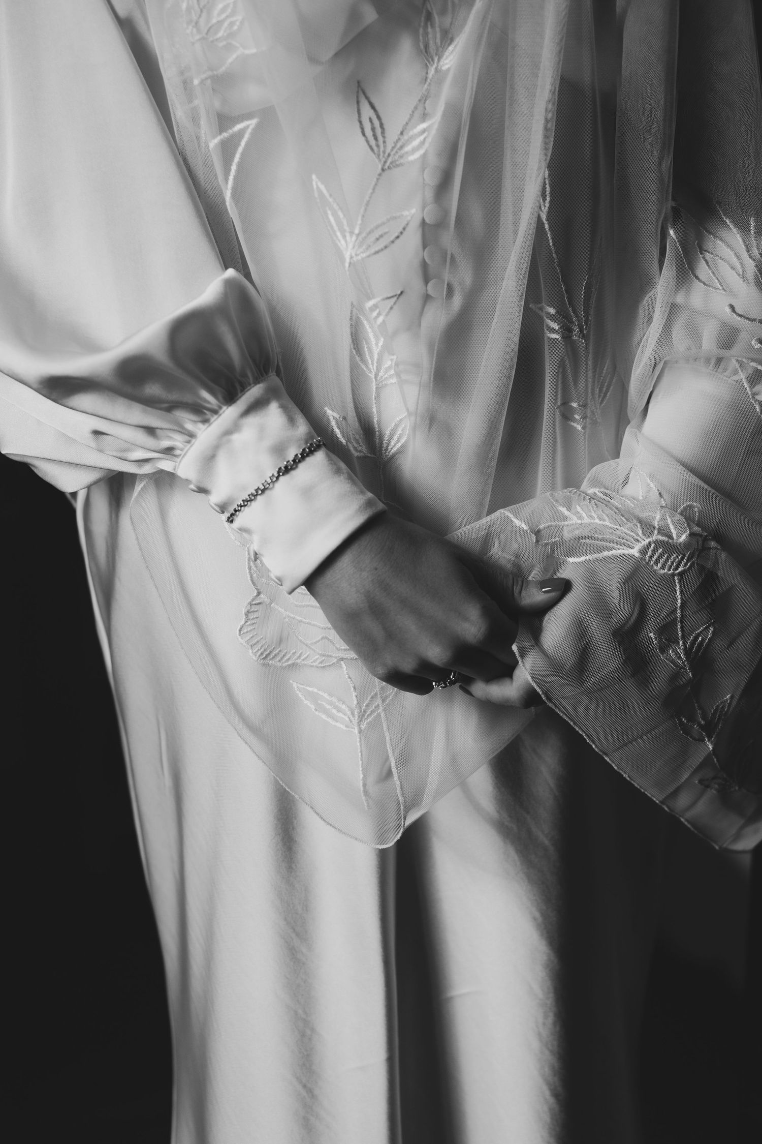 Black and white close-up of hands adjusting the sleeve of an elegant white robe with delicate floral embroidery.