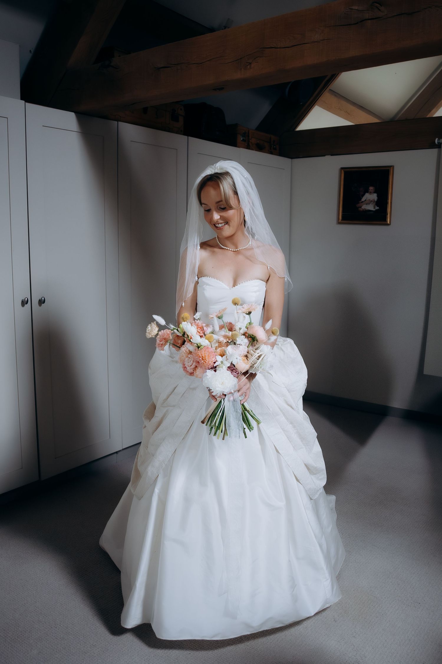 A bride in a white wedding dress holds her wedding bouquet of pink and peach flowers at bridal preparations.