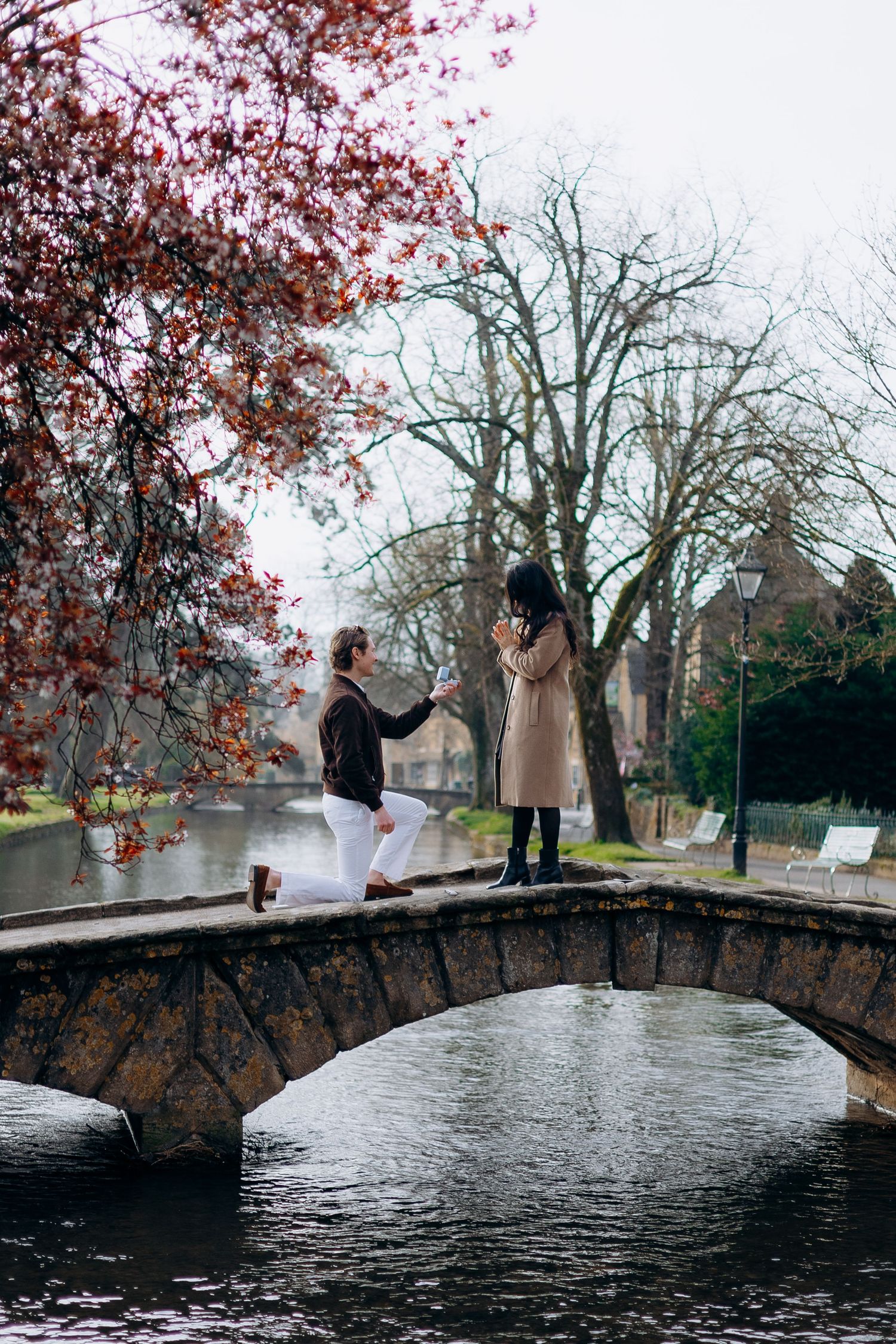 Man proposes in the cotswolds over a river in autumn, with a woman standing before him in a romantic outdoor setting.