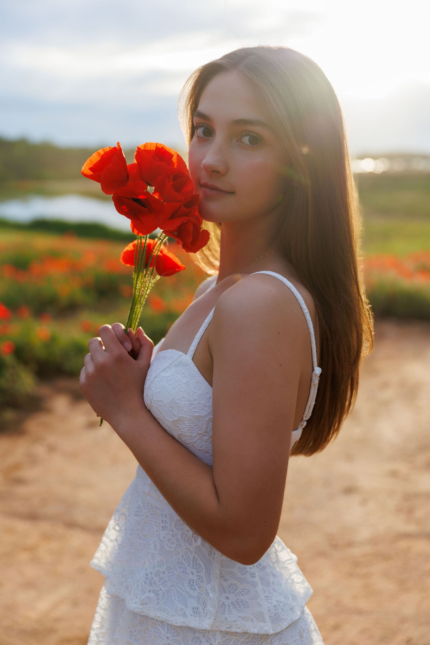 A person in a white lace dress poses with red poppies in a field at sunset.
