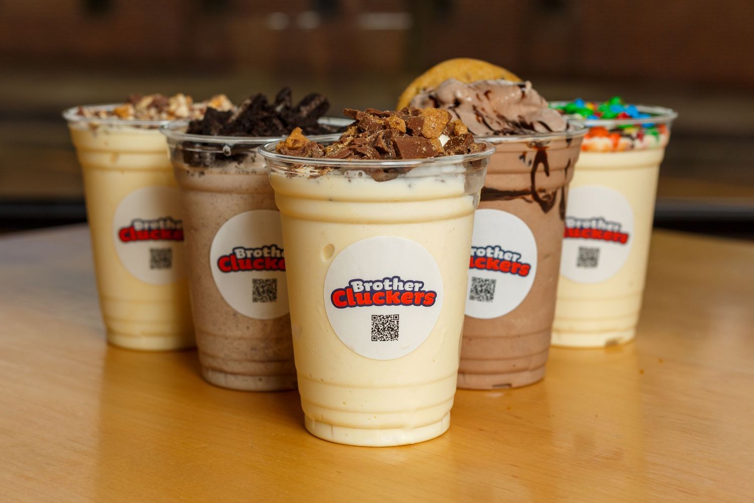 Five creamy milkshakes with toppings arranged on a wooden table with Boston Burger Company branded cups.