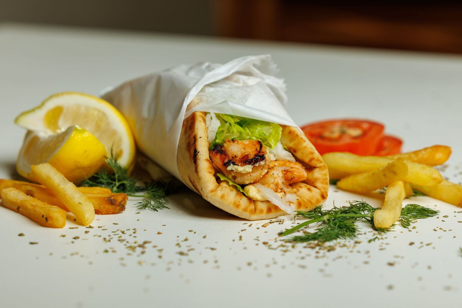 Grilled chicken shawarma wrap served with french fries, lemon wedge and fresh herbs on white plate.