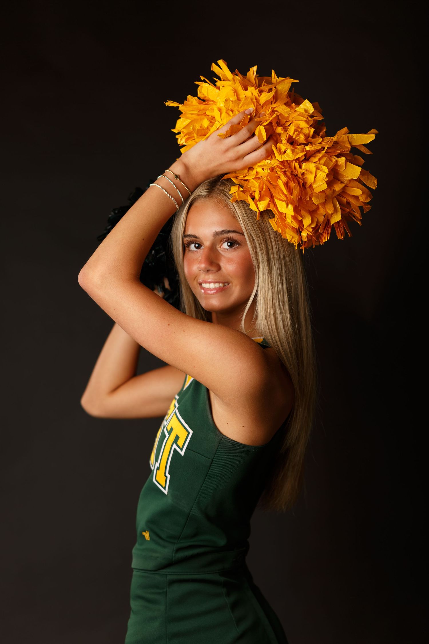 Cheerleader in green uniform poses with yellow flower crown against dark background.
