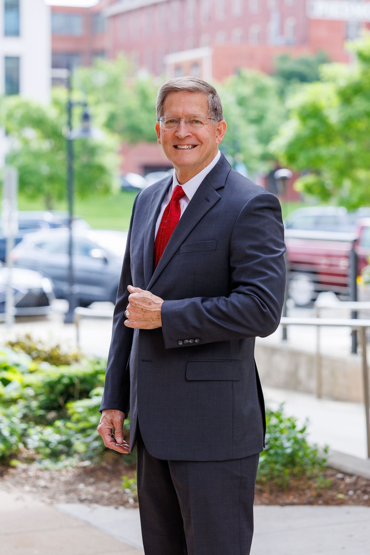 Professional portrait of someone in a dark business suit with red tie standing outdoors near landscaping and buildings.
