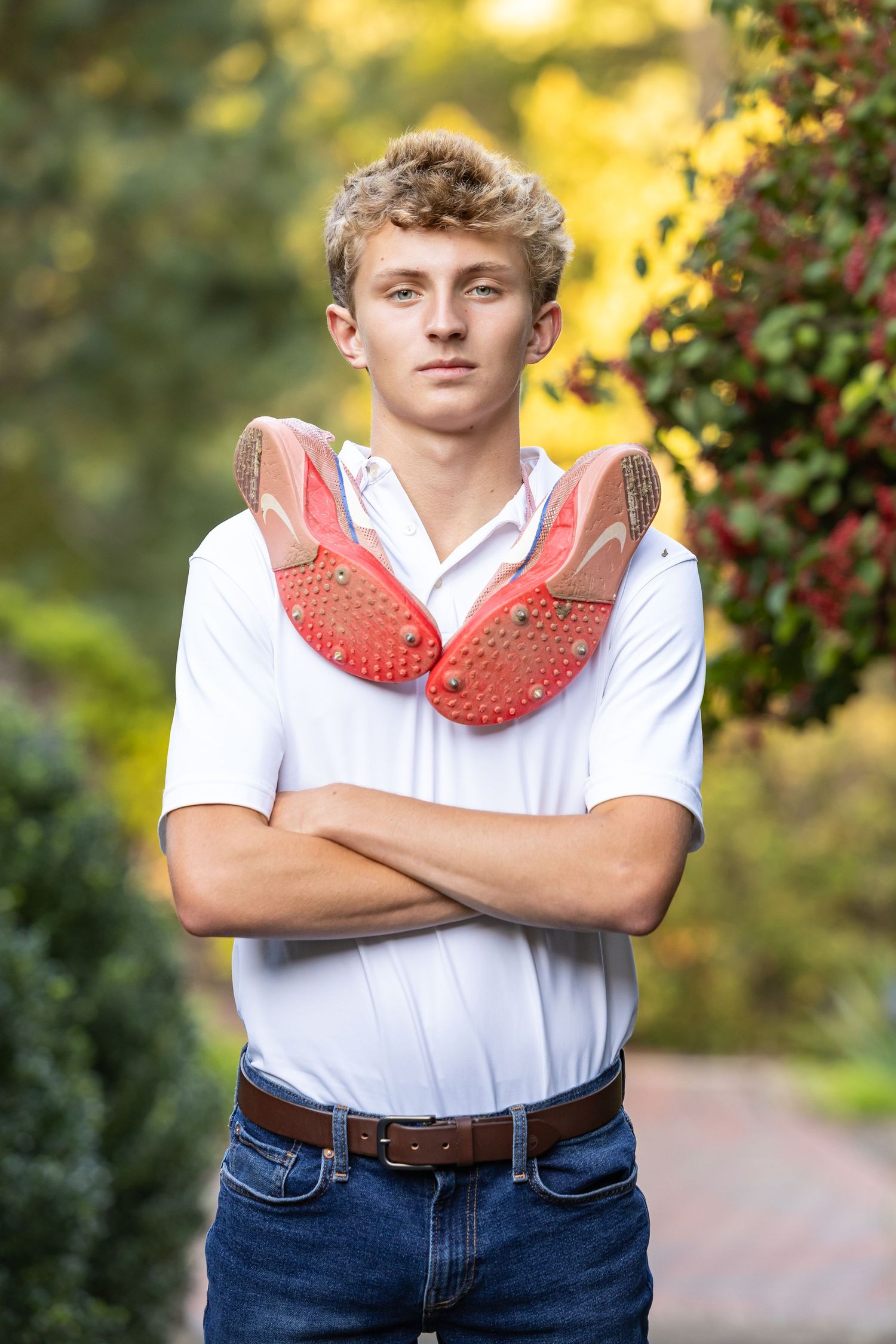 Young person in white polo shirt and jeans poses outdoors with red sports cleats draped over crossed arms.