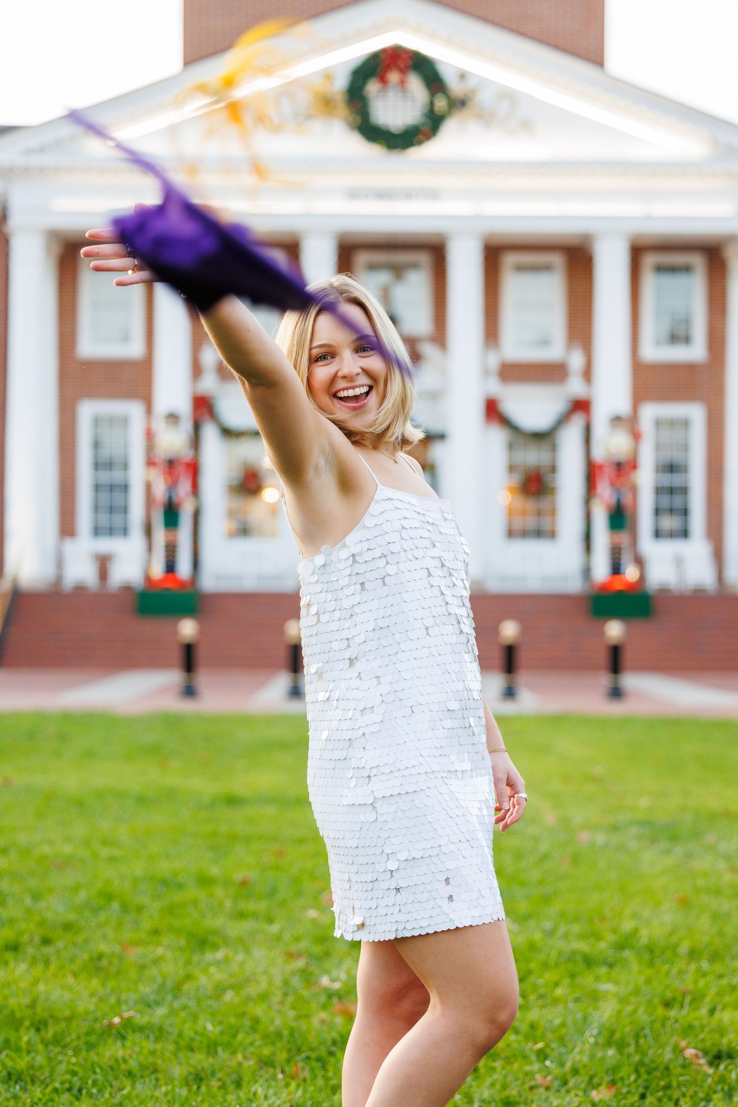 A graduate celebrates on a lawn in front of a brick building decorated with holiday wreaths and garland.