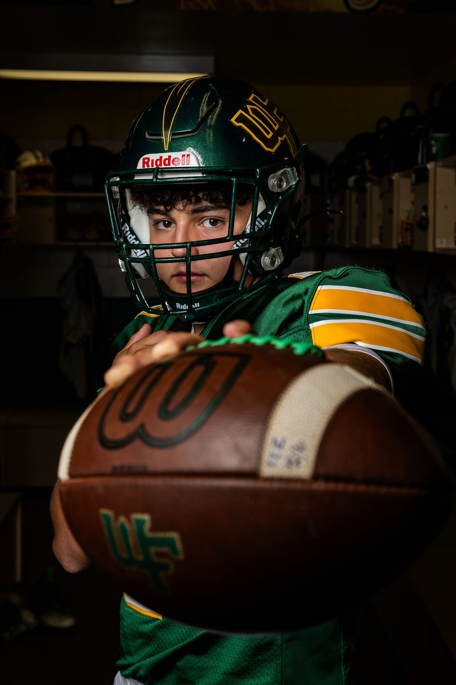 A football player in green and gold uniform holds out a Wilson football in a dark locker room setting.