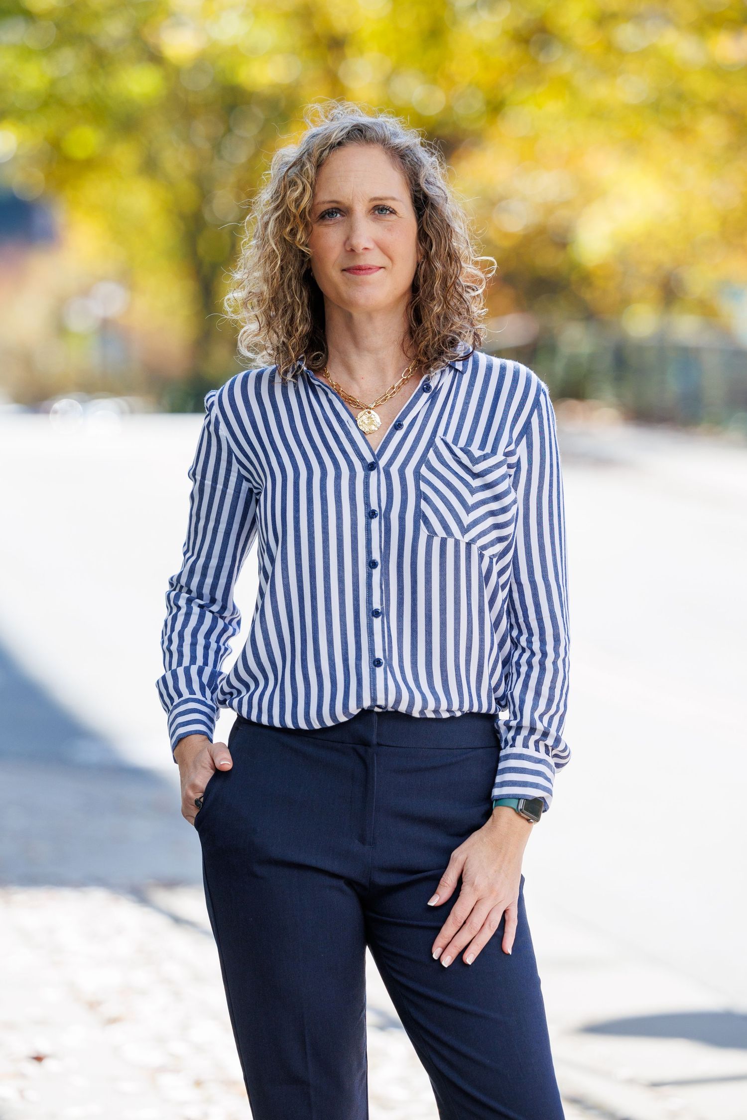 Professional portrait outdoors showing a person in a blue striped button-down shirt and navy pants against autumn foliage.