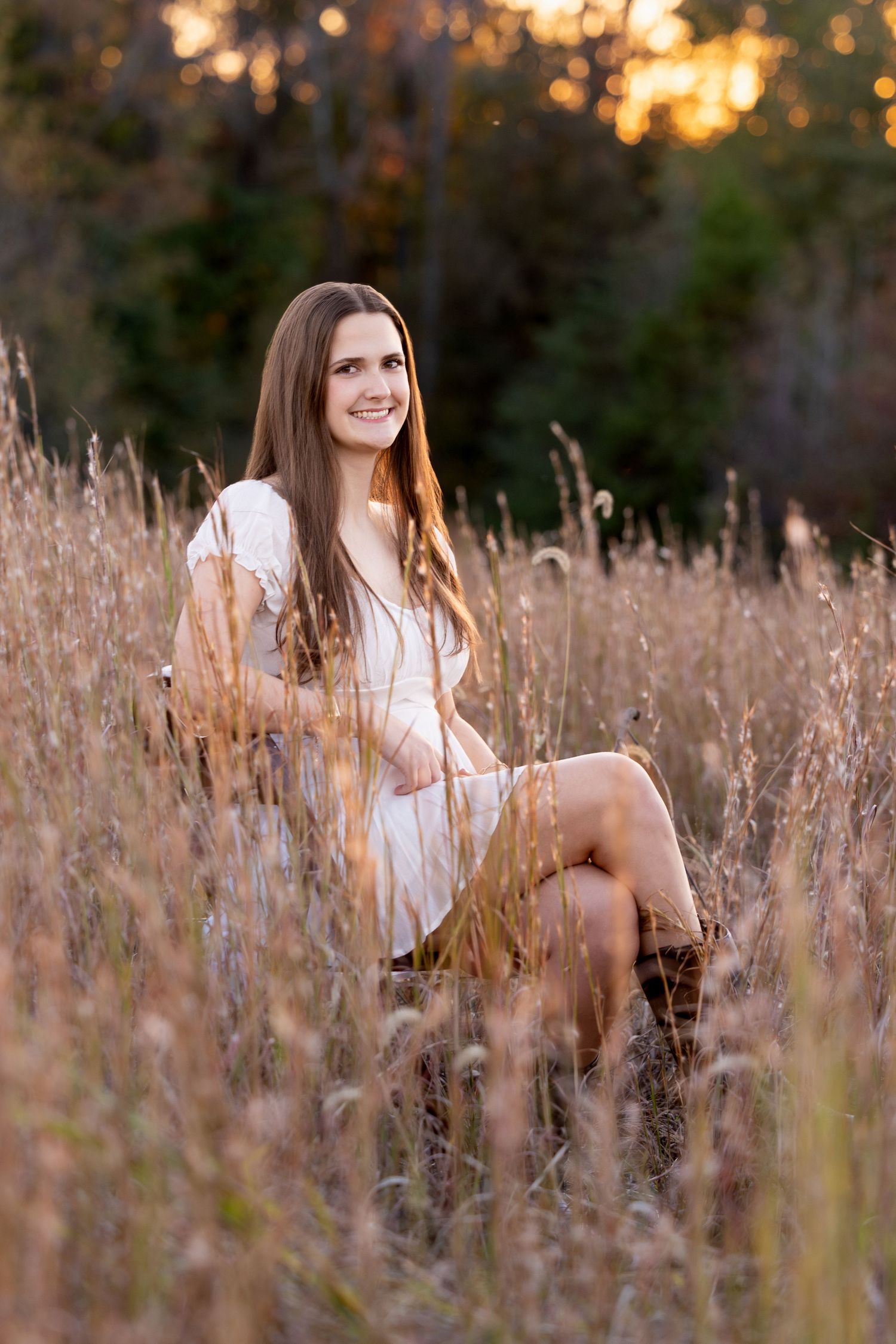 A woman in a white dress sits gracefully in a golden field of tall grass during sunset.