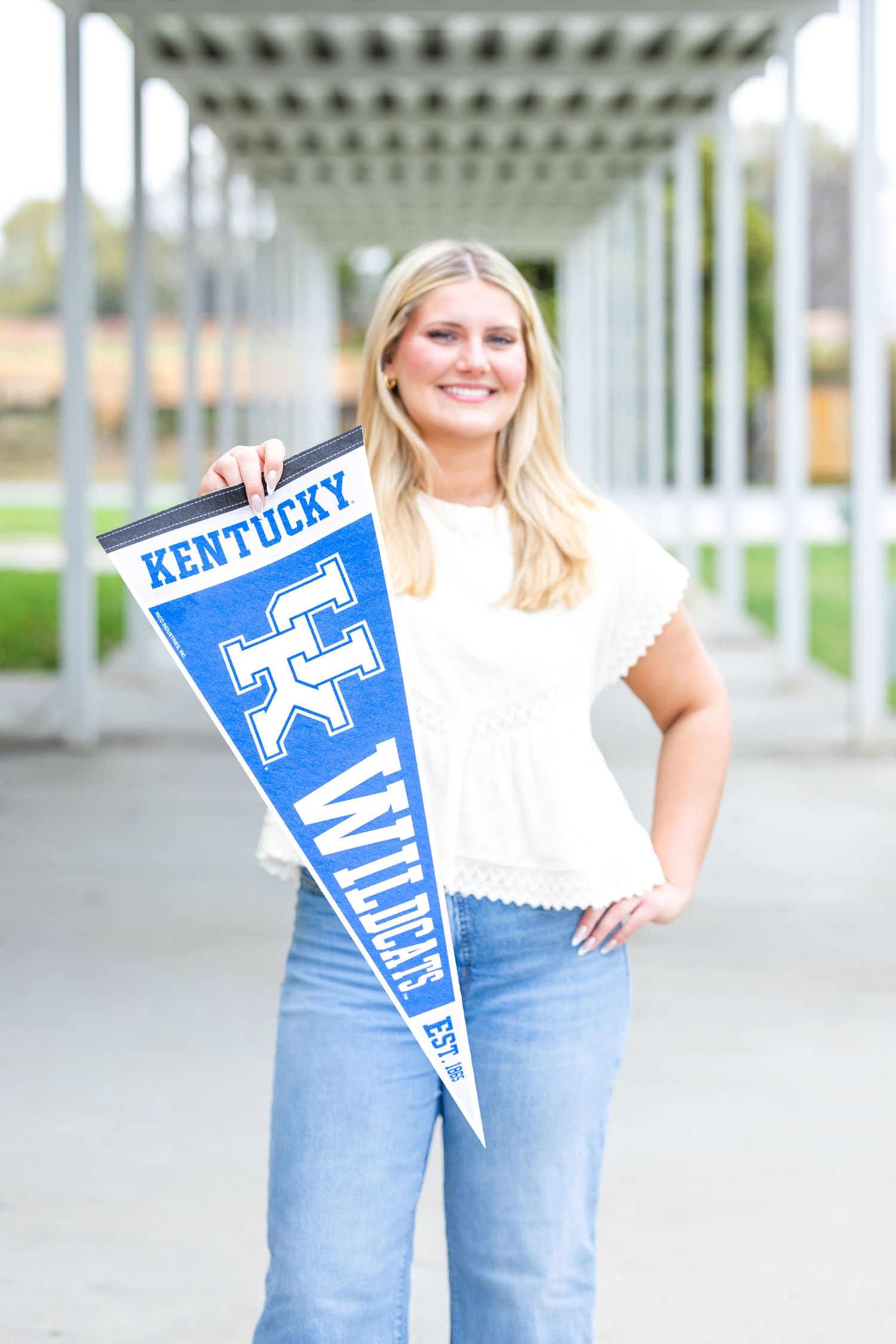 A person in a white top and jeans holds a blue University of Kentucky pennant flag in an outdoor setting.