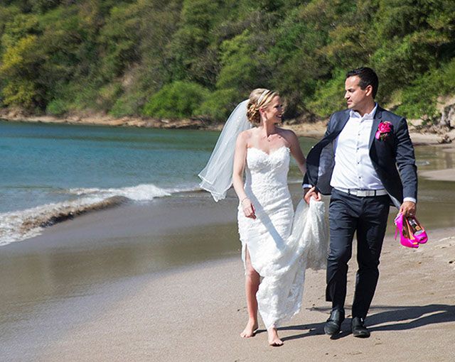 A couple strolls on a beautiful beach during their destination wedding with turquoise waters and lush greenery behind them.