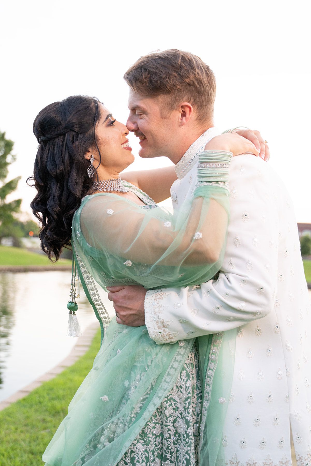 A romantic couple shares an embrace outdoors, with the woman wearing an elegant mint green traditional Indian lehenga.