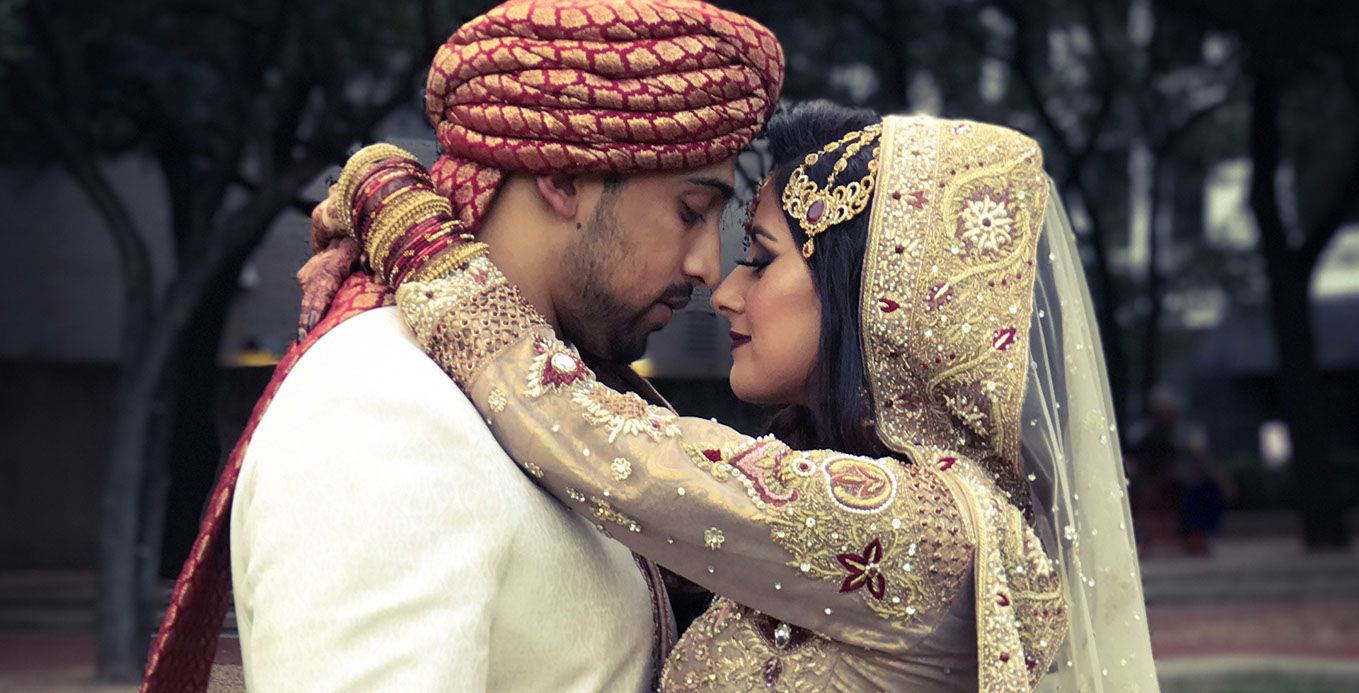 A stylish South Asian couple wearing traditional wedding attire in cream and gold share an intimate moment outdoors.