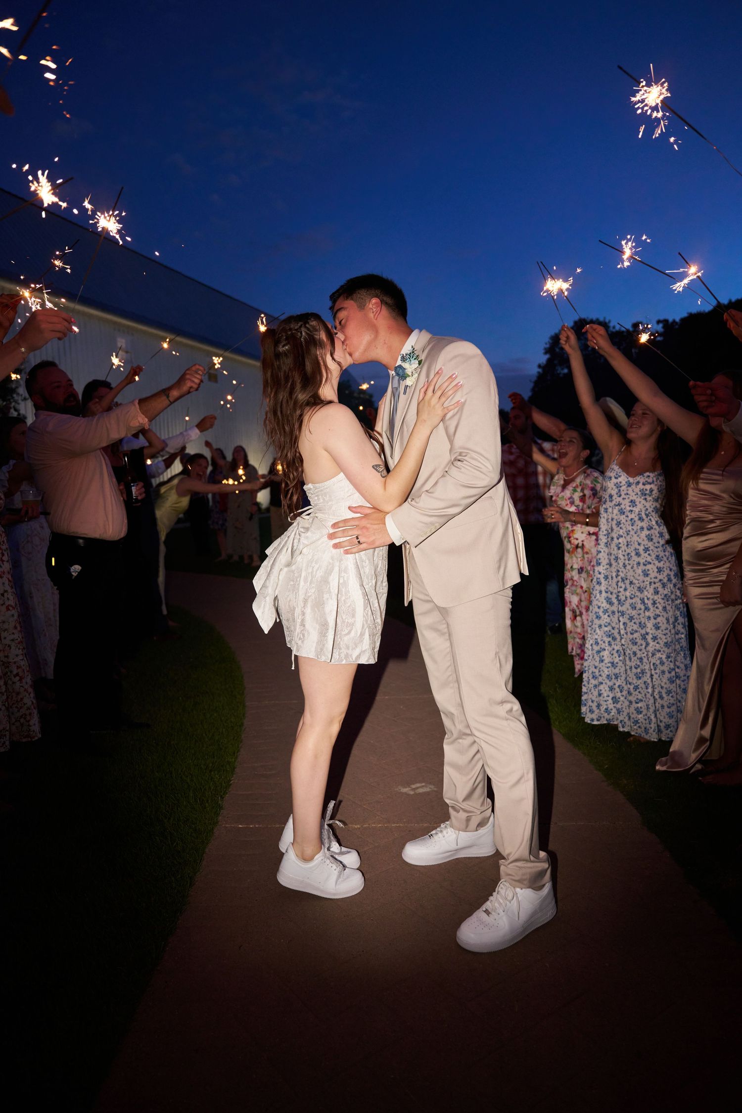 A couple shares a romantic kiss under sparklers at an evening outdoor celebration while guests cheer around them.