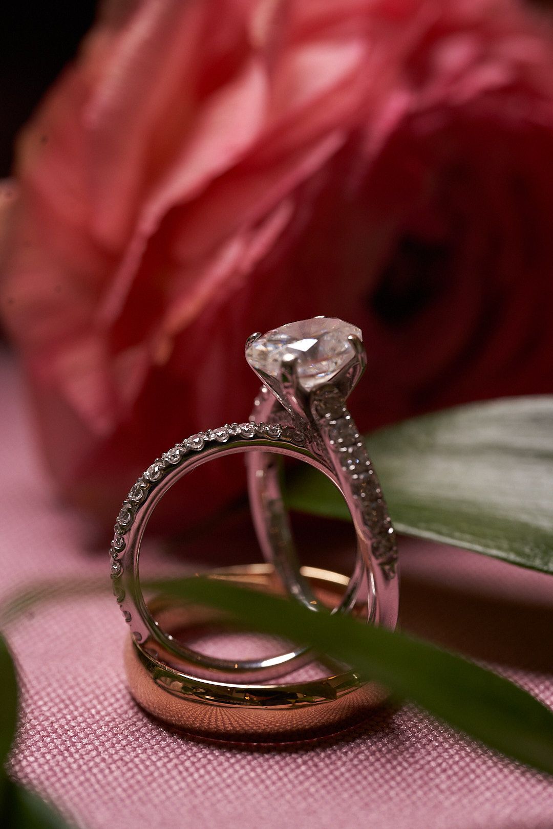 Diamond engagement ring and wedding band displayed with a pink rose and green leaf on soft fabric.