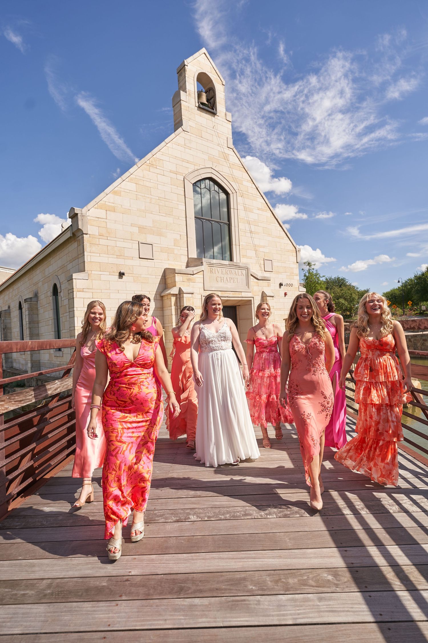 Bridal party in floral coral dresses walking along wooden boardwalk outside stone church with bell tower on sunny day.
