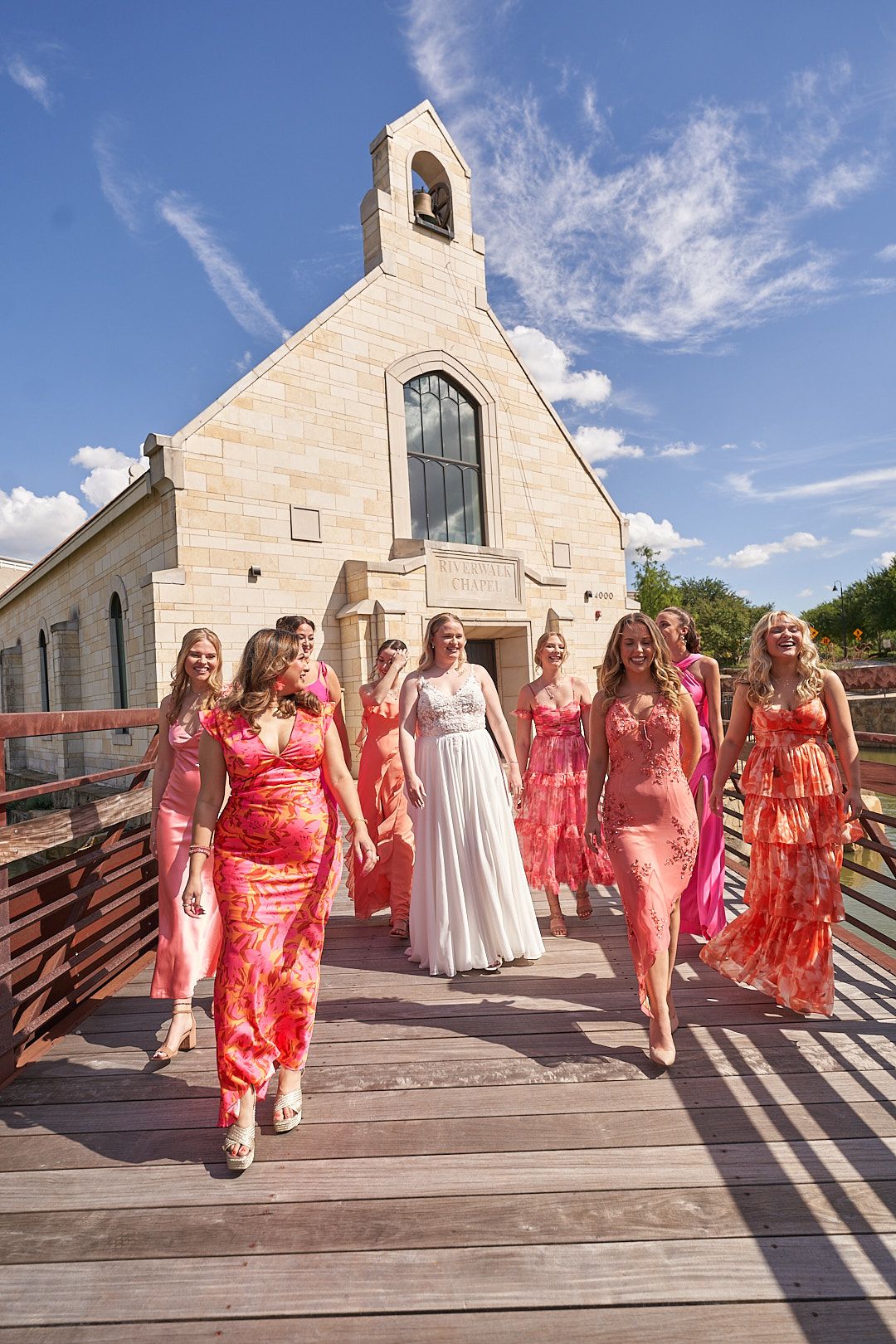 Wedding party in coral dresses walking outside stone church under blue sky.