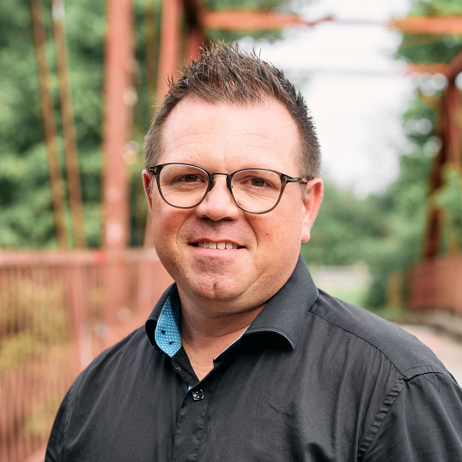Professional headshot against blurred green and brown outdoor backdrop wearing glasses and black shirt.