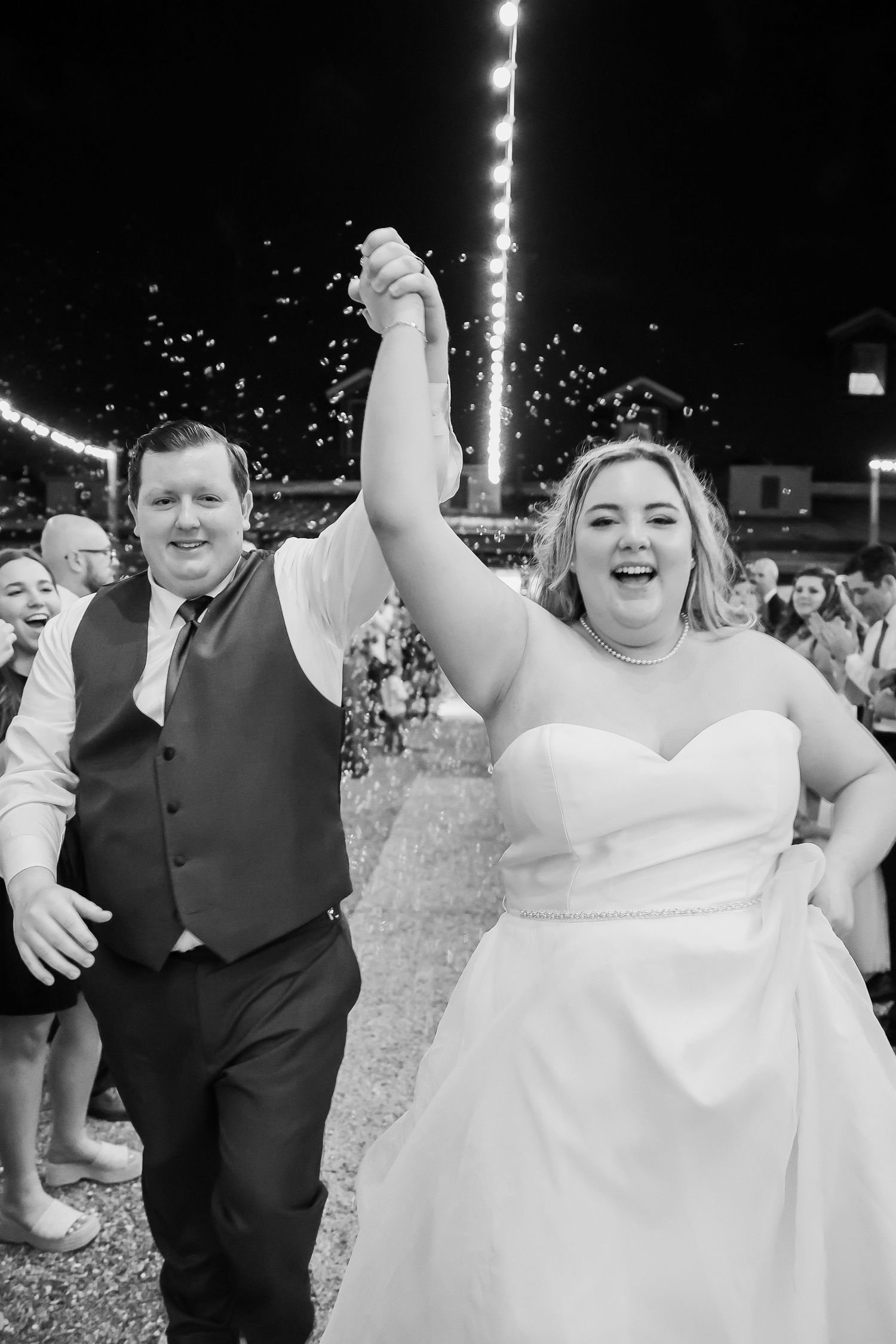 A joyful black and white photo of newlyweds celebrating at their wedding reception with string lights in the background.