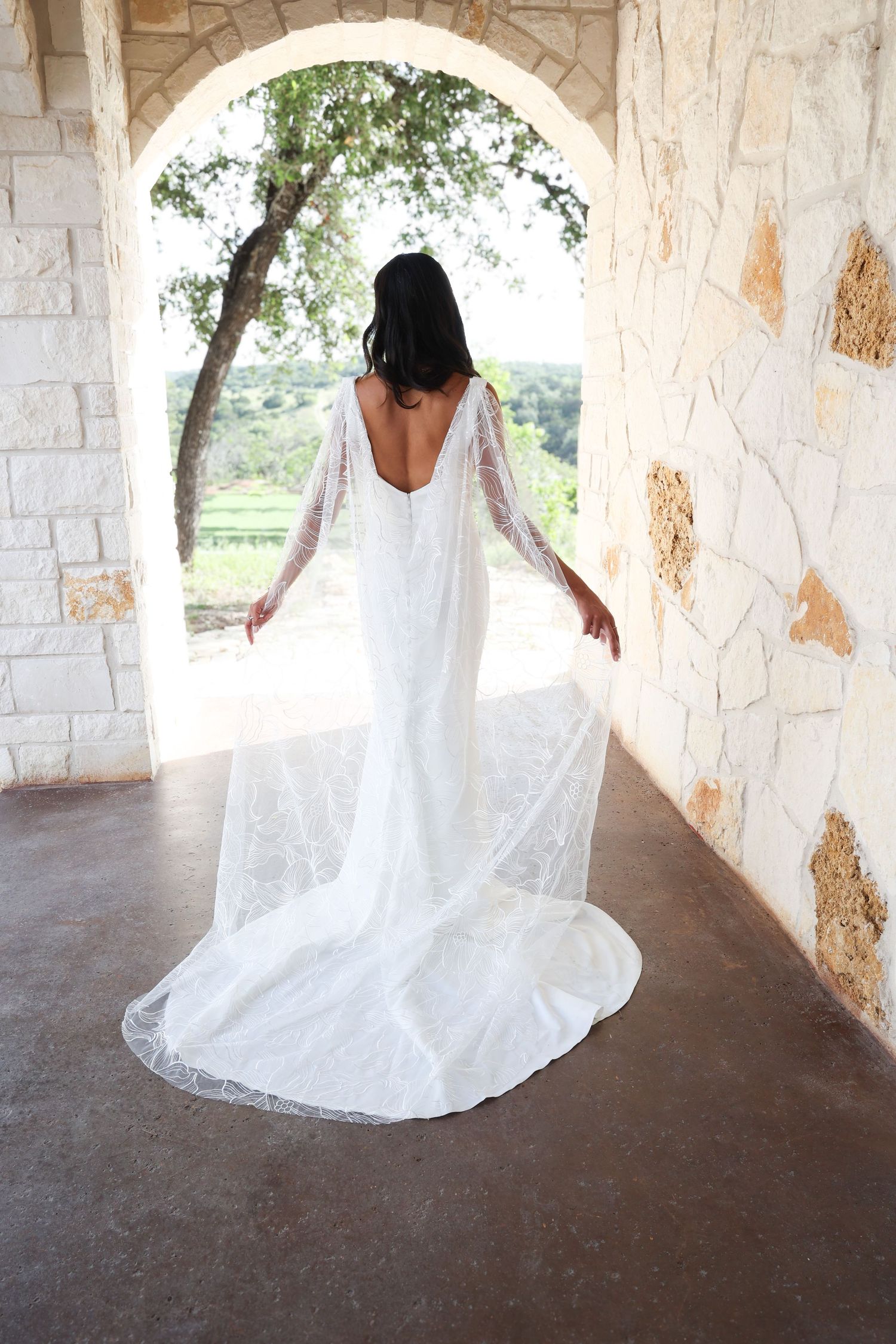 Back view of bride in backless wedding gown standing under stone archway.