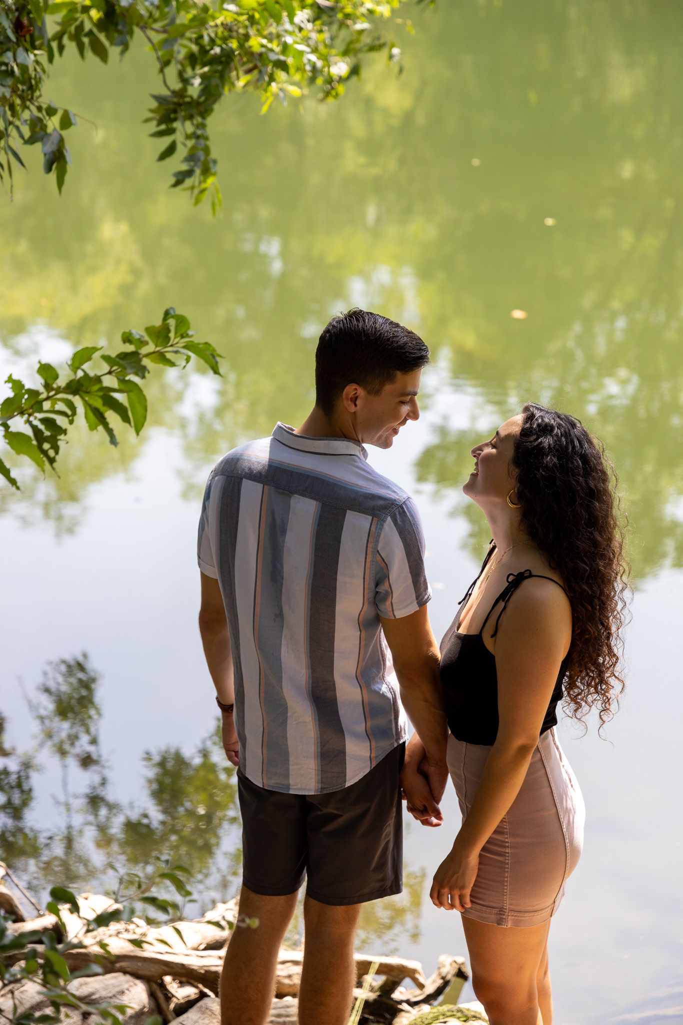 A couple stands together beside a tranquil pond on a sunny day, holding hands amidst lush greenery at Red Bud Isle in Austin, TX.
