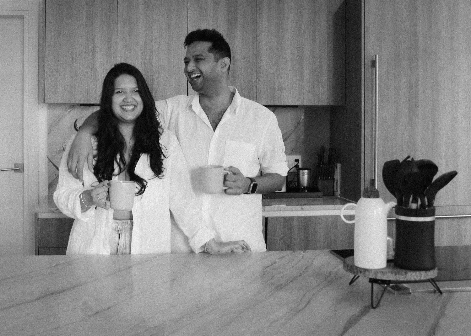 A couple in white shirts share a morning moment in a minimalist kitchen with wooden countertops and coffee essentials in Miami, FL.