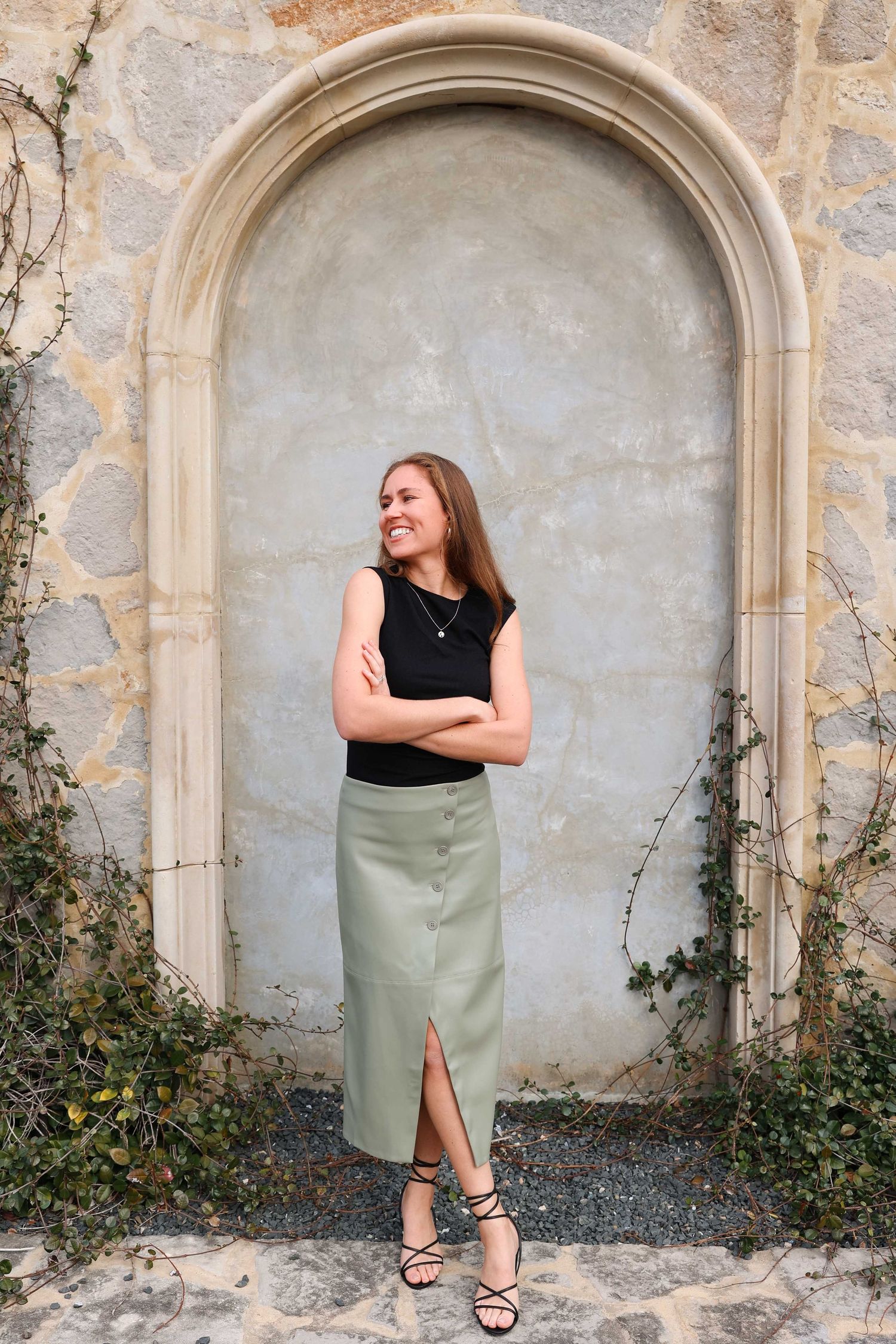Person in black top and sage green skirt poses in stone archway with climbing vines.