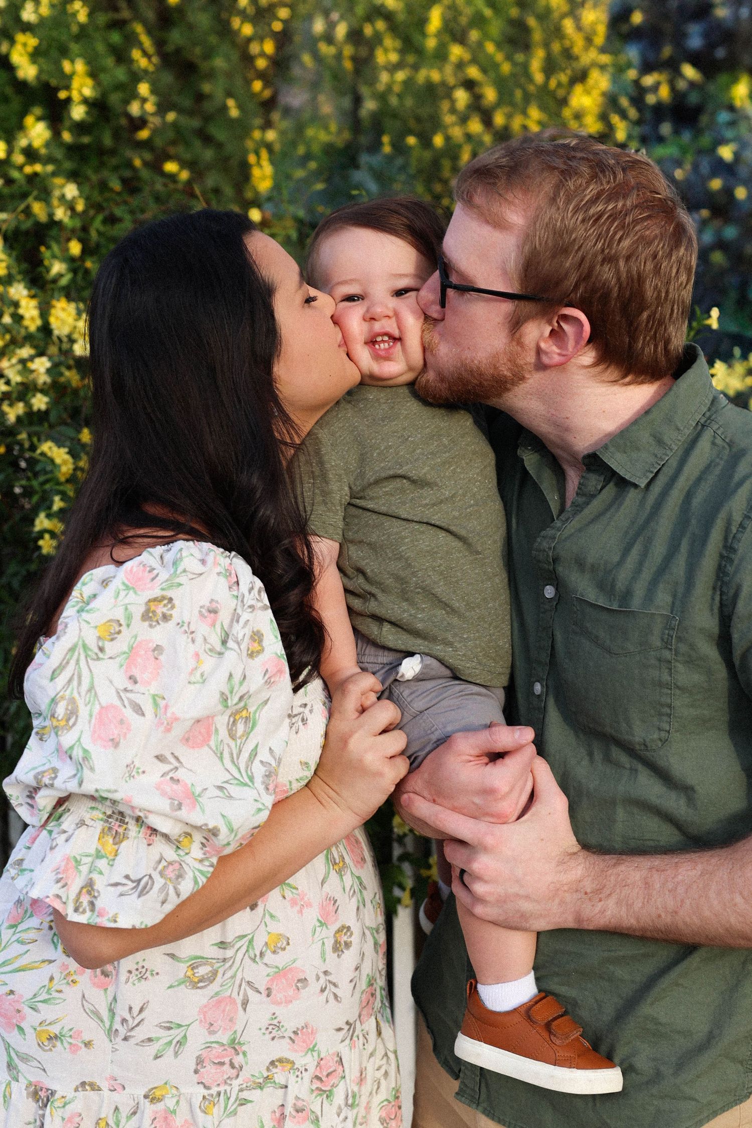A couple and baby share sweet kisses together outdoors against a backdrop of blooming yellow flowers.