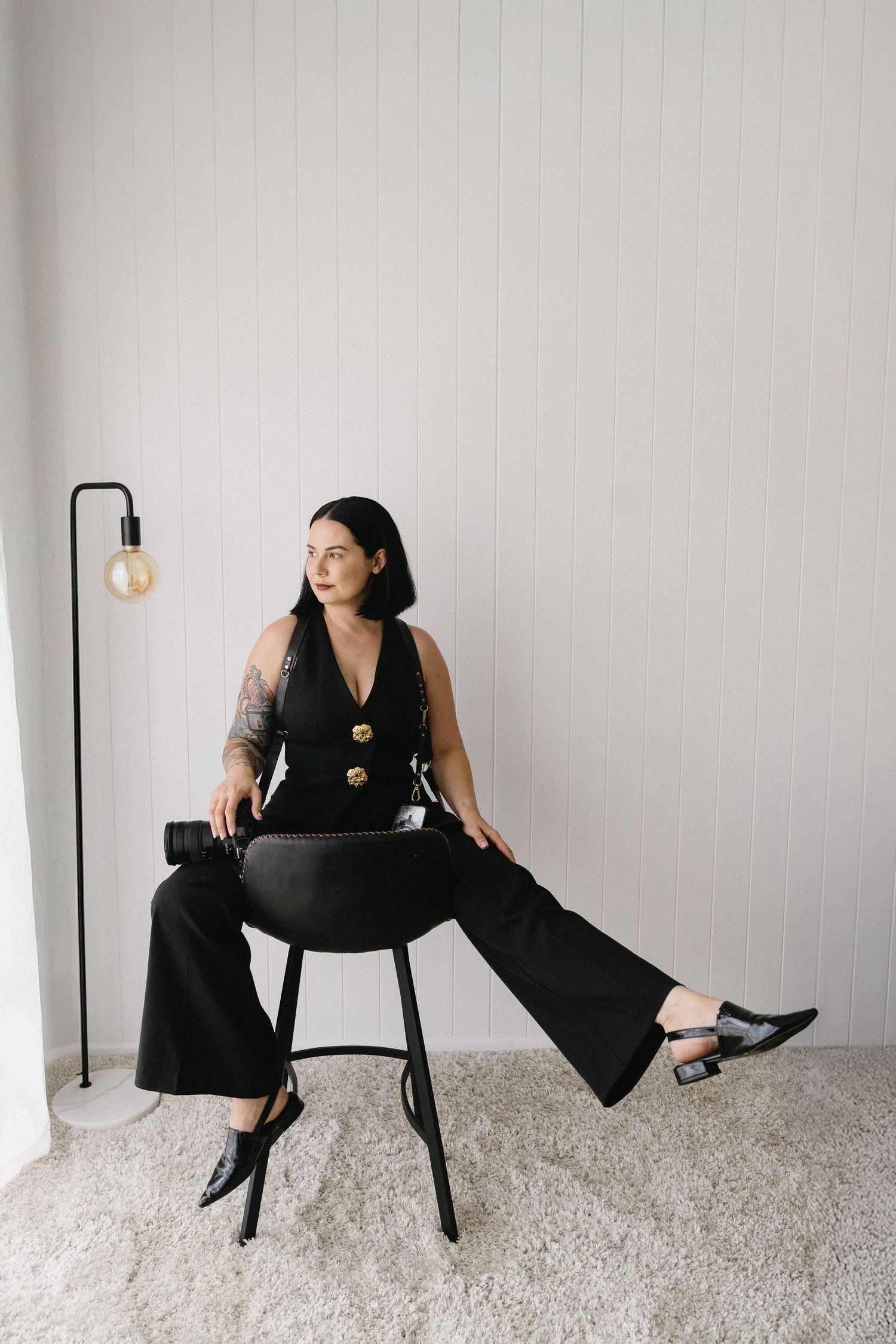A person in black attire poses artistically on a chair against a white wall in a minimalist setting.