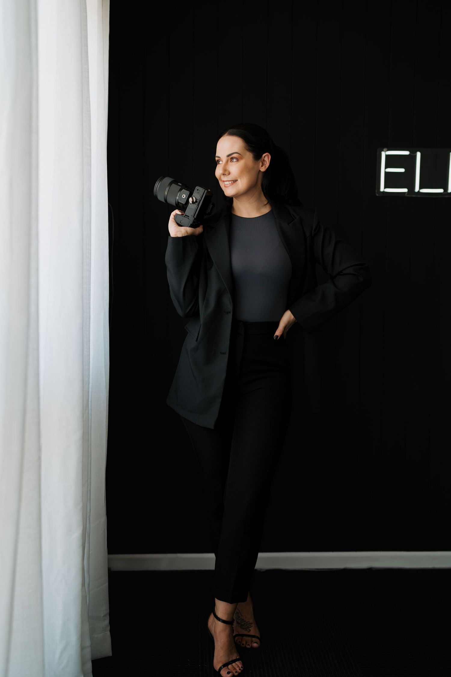 Elegant photographer in black outfit poses with camera against dark wall.