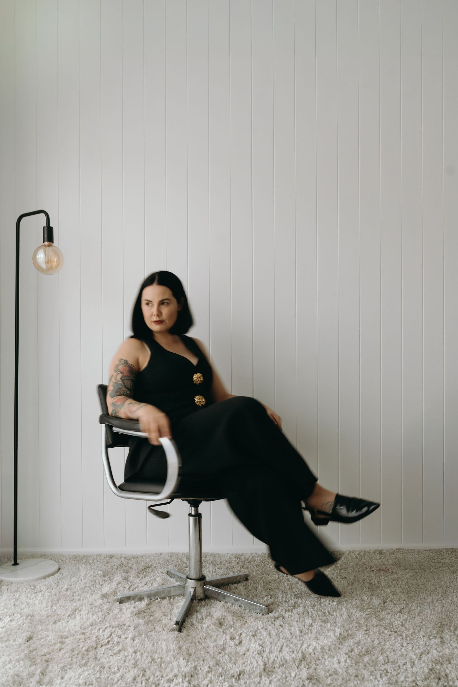 A figure in a black dress poses dramatically in different positions on a white chair against a minimalist gray wall.