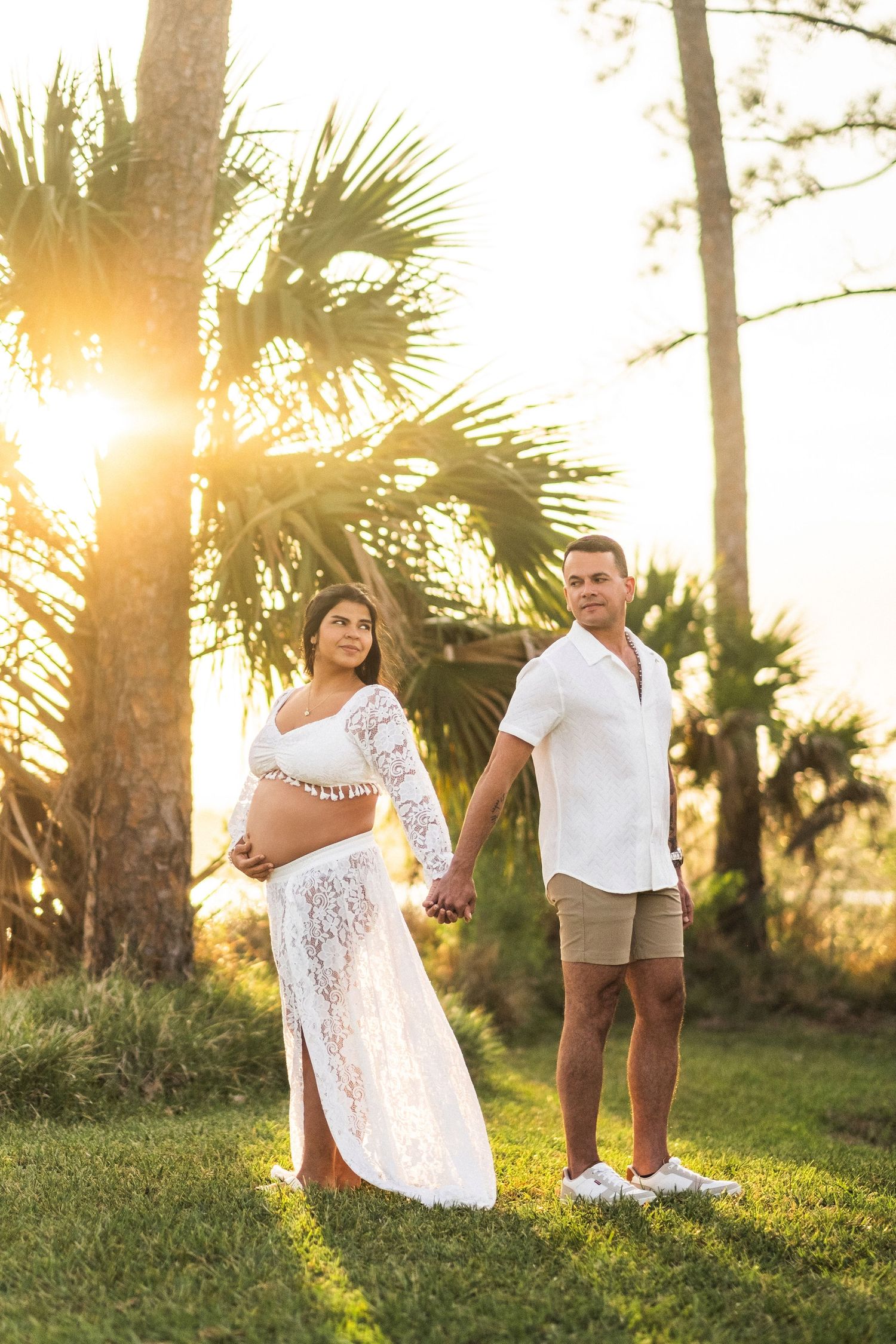 Pregnant couple in white outfits hold hands at golden sunset among tropical palm trees.