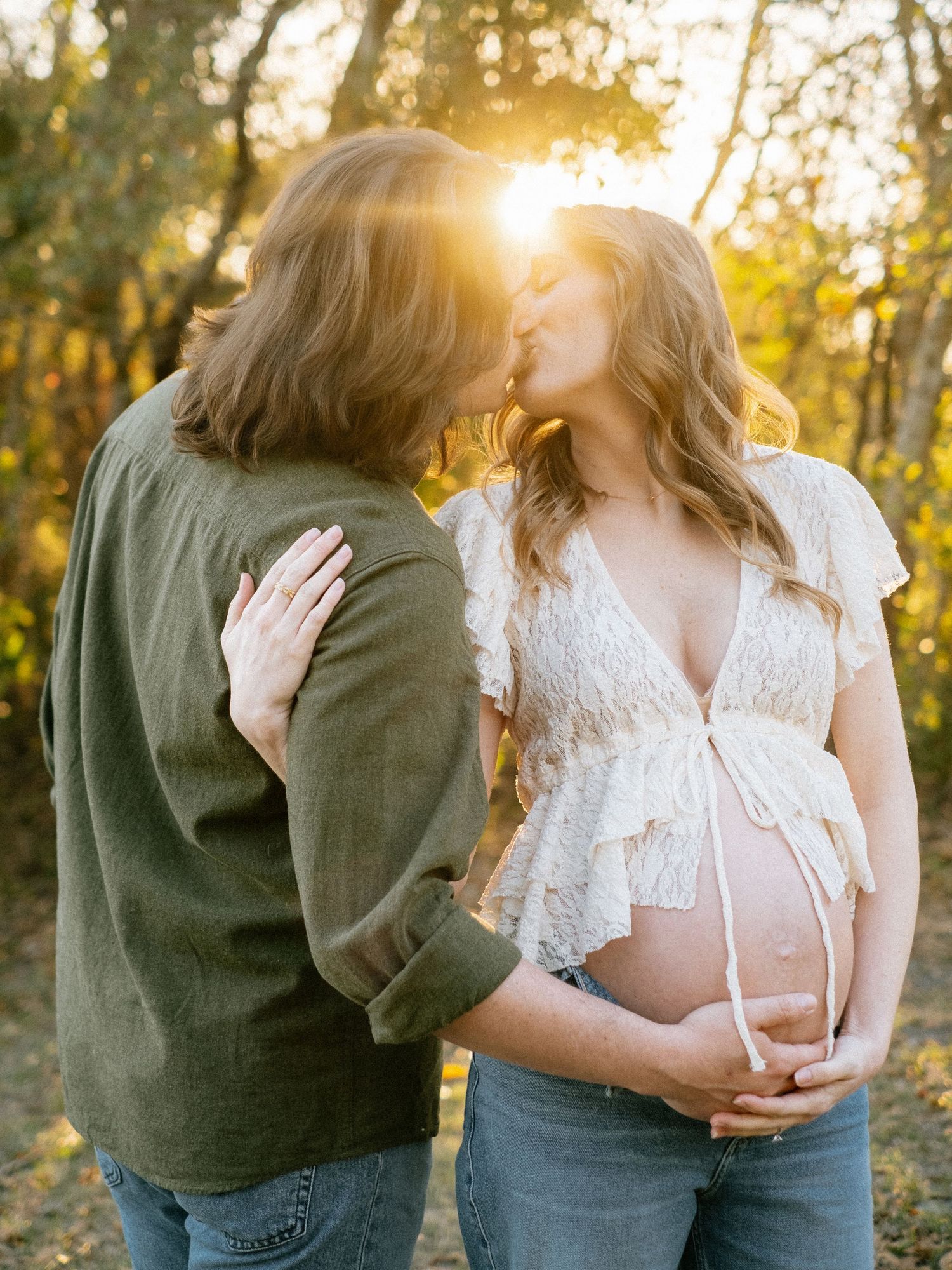 Couple kissing at golden hour in autumn woods, pregnant woman in lace top holding baby bump.