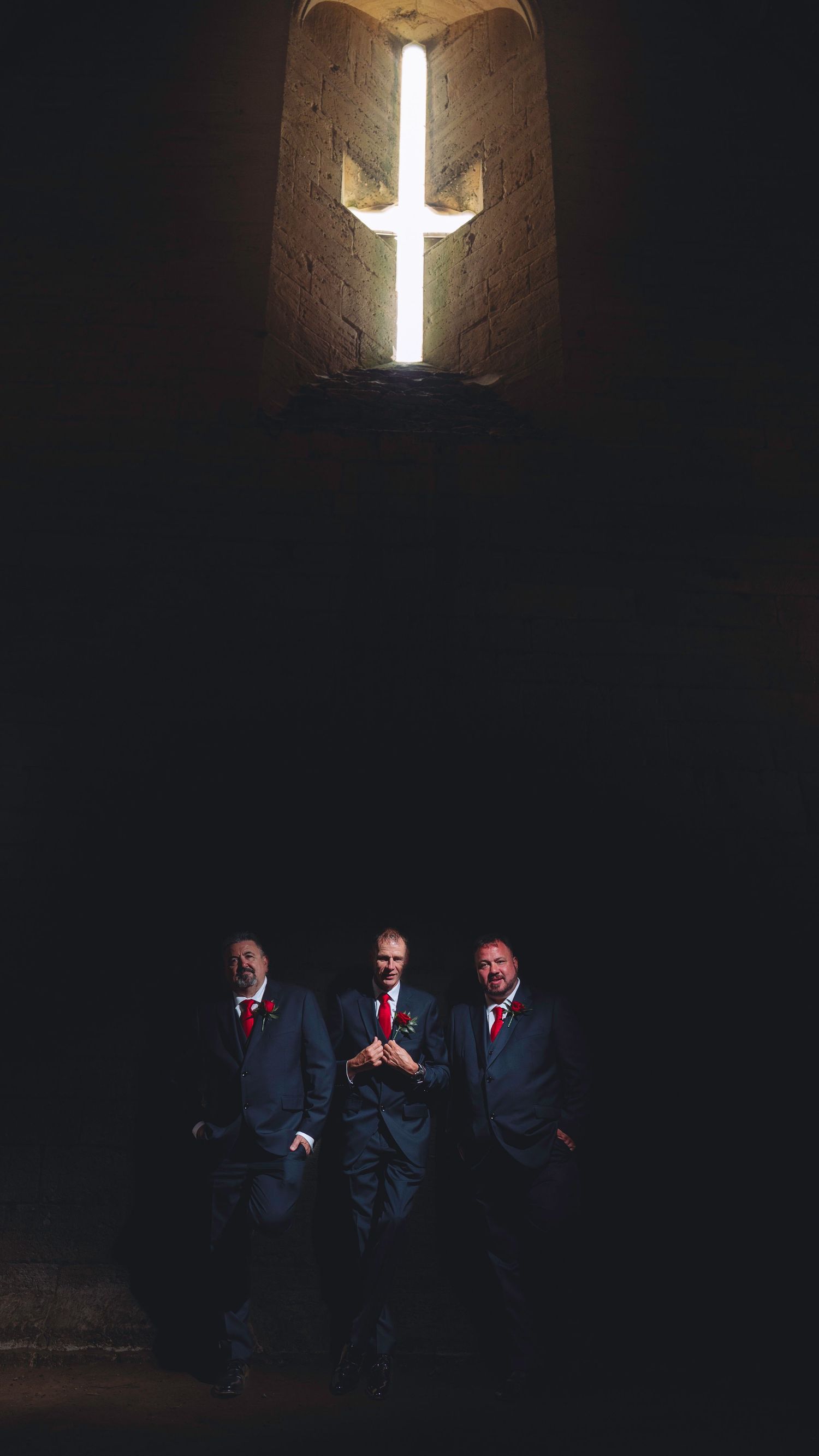 Three figures in dark suits and red ties stand beneath a glowing cross-shaped window in a dimly lit stone chamber.