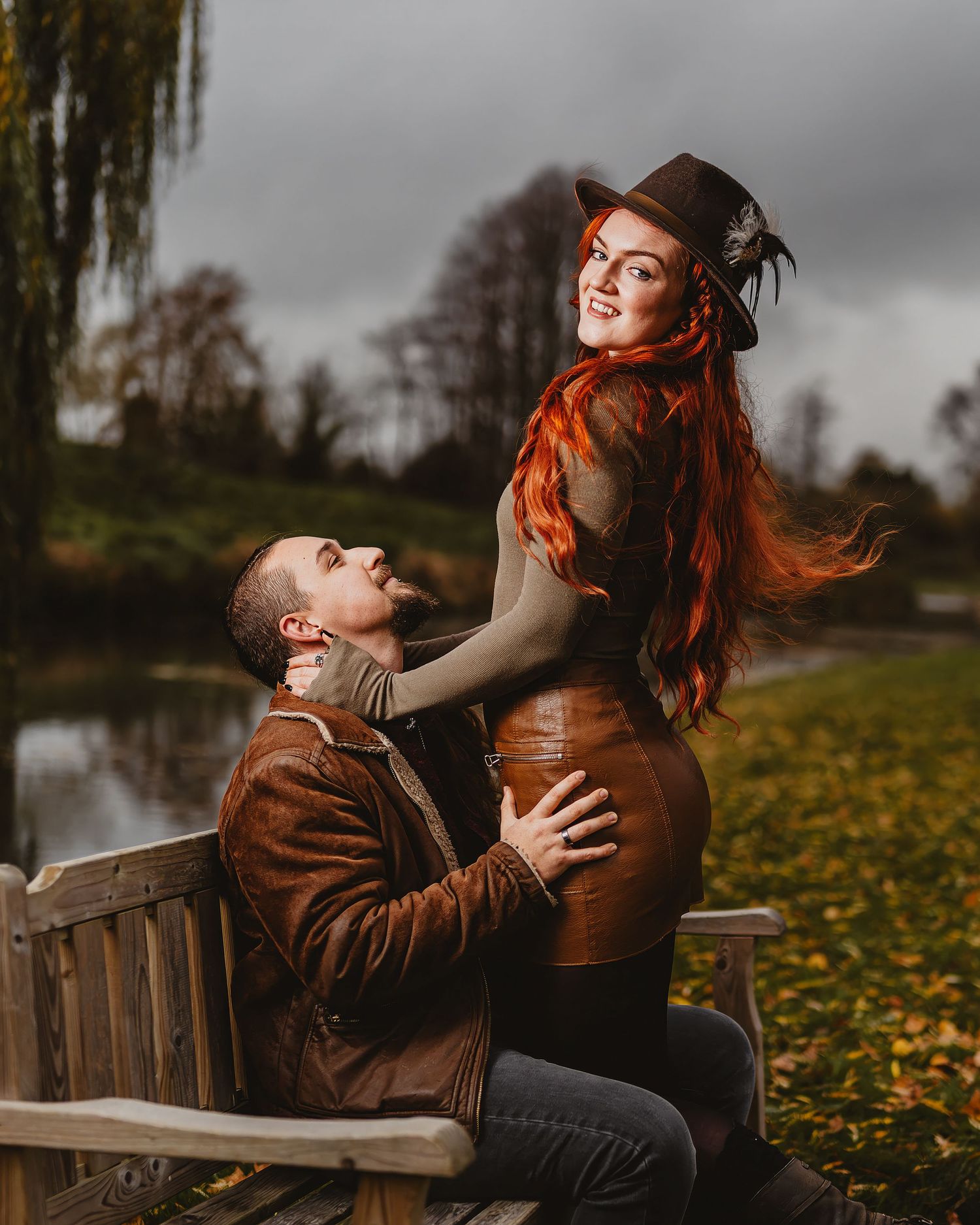 A couple shares tender moments on a wooden bench surrounded by fallen autumn leaves on a moody overcast day.