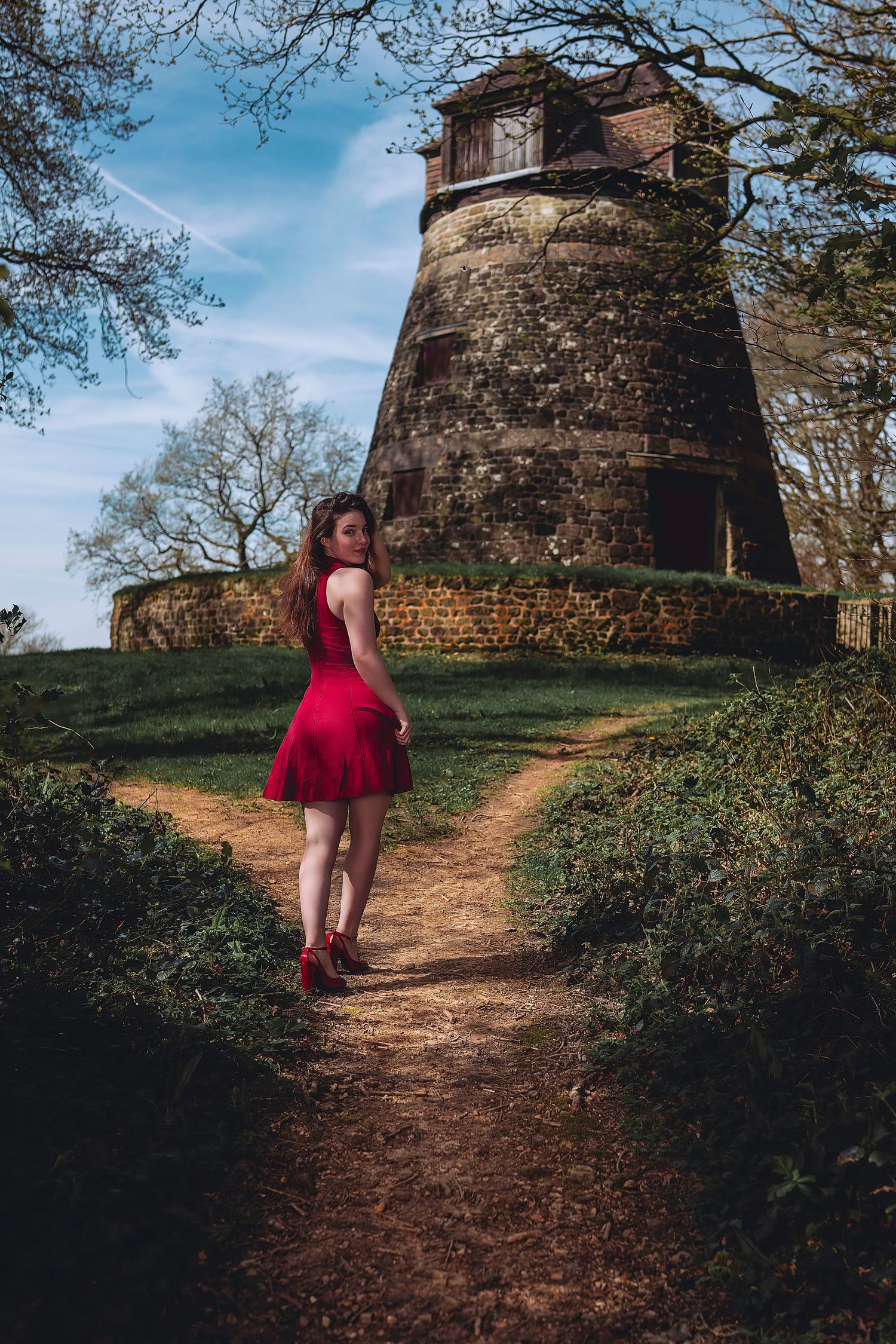 A red dress contrasts against an ancient stone tower on a winding dirt path through a scenic woodland landscape.