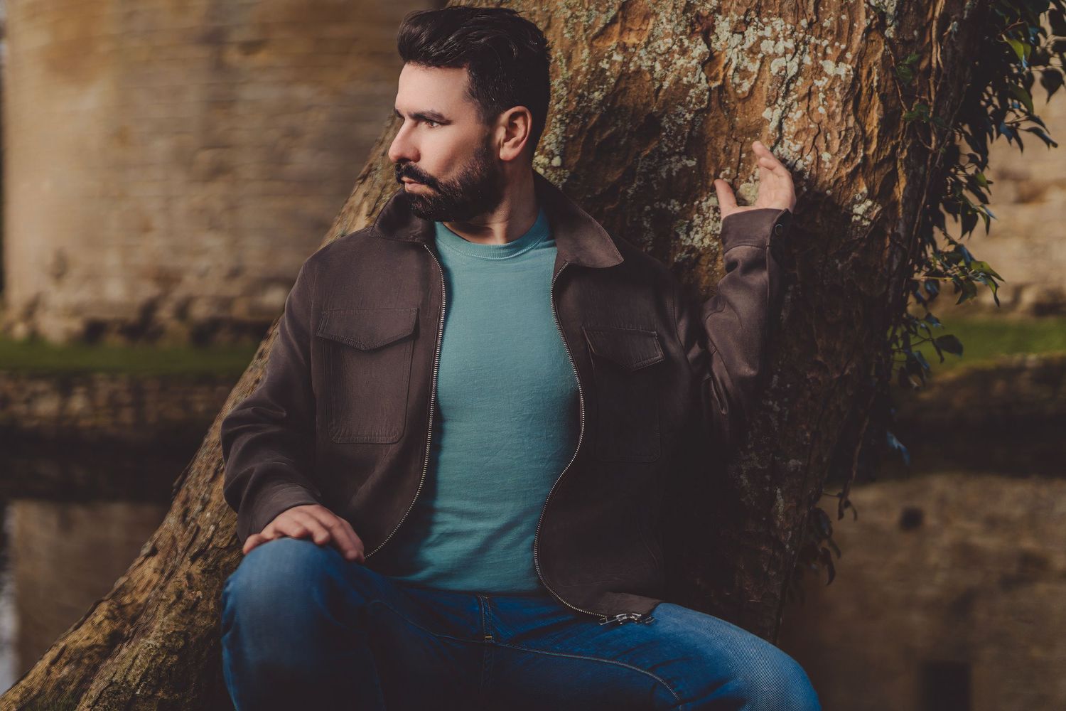 Person wearing brown jacket and blue shirt sits on stone wall in outdoor setting.