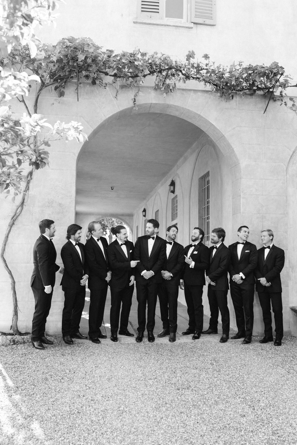 Groom and groomsmen in classic black tuxedos posing together under an arched stone villa entrance in black and white.
