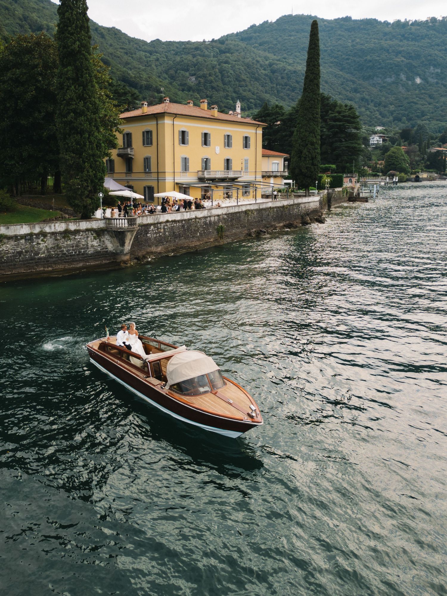 Couple riding a classic wooden speedboat on Lake Como with a yellow Italian villa and lush green hills in the background.