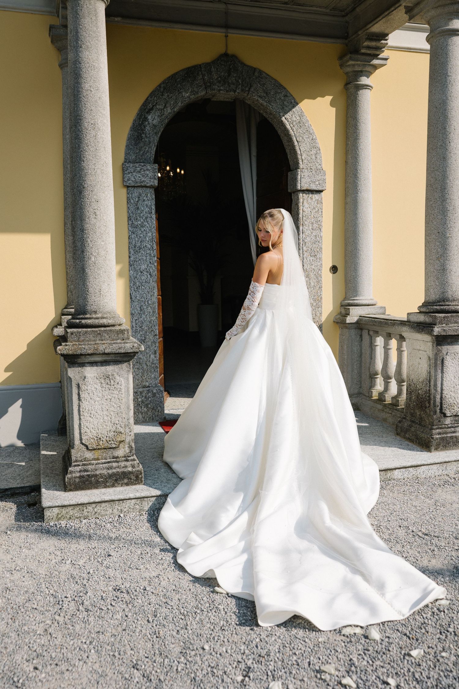 Bride in elegant white ball gown with long train stands at entrance of classic stone villa with arched doorway.