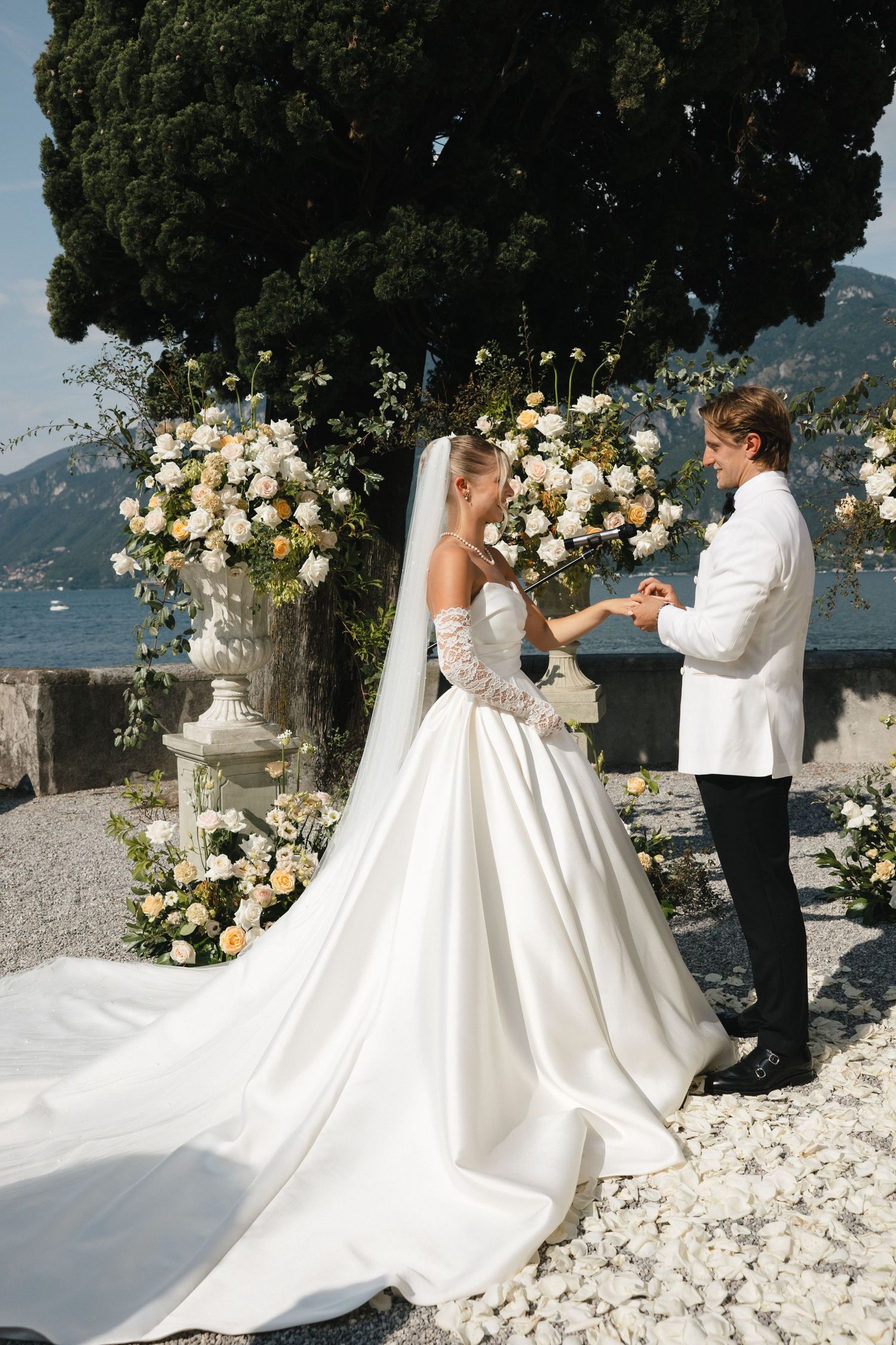 Bride in white ball gown and groom in white jacket exchange rings at outdoor wedding ceremony with floral arrangements.