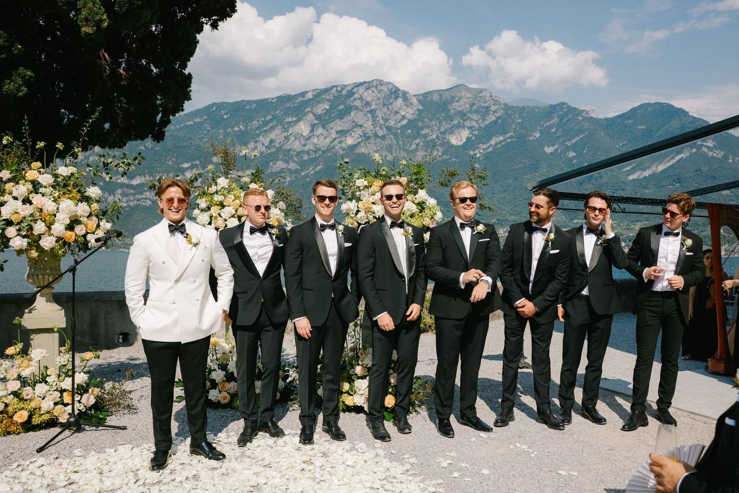 Groom and groomsmen in black tuxedos pose at an outdoor wedding ceremony with mountain and lake views.