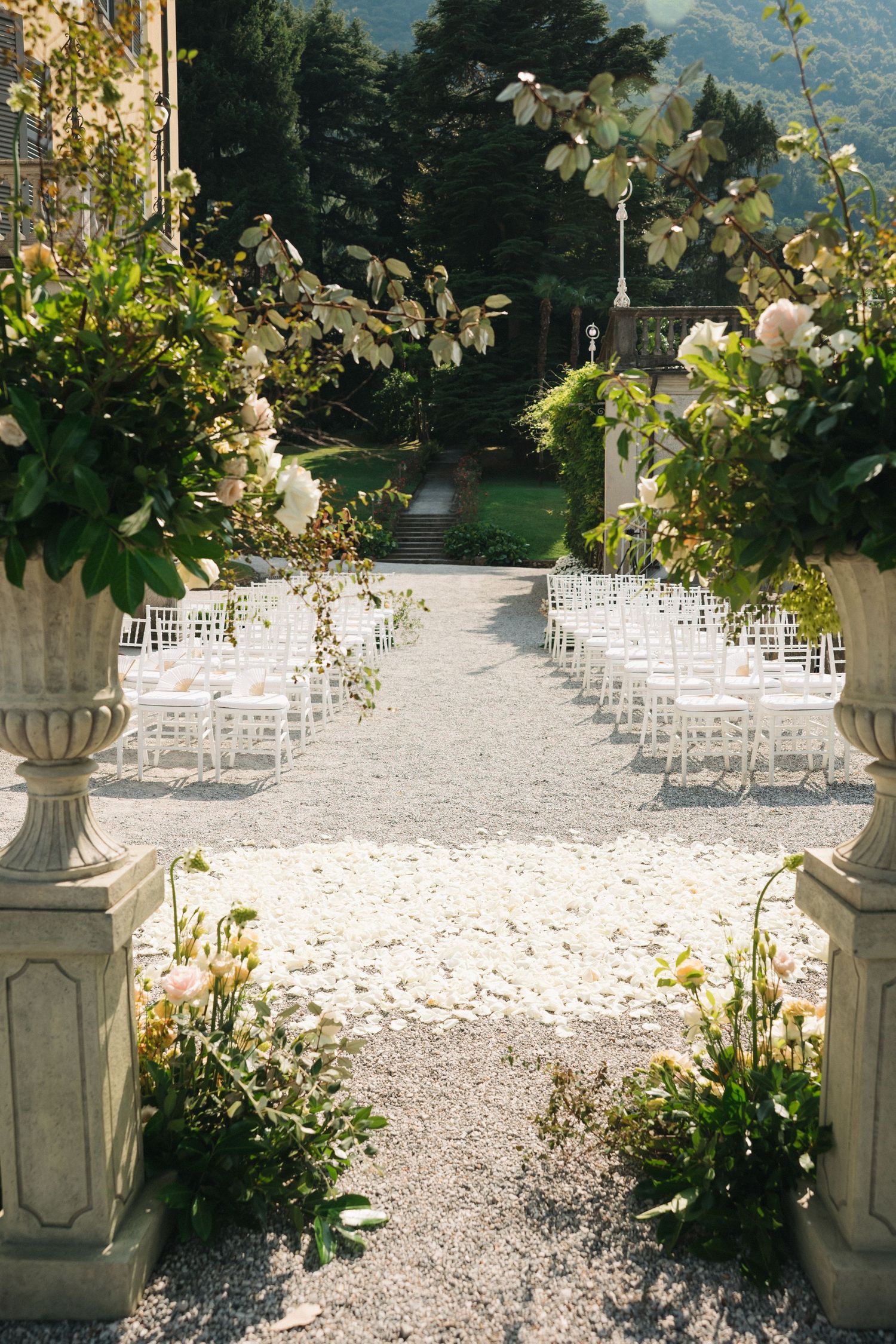 Elegant outdoor wedding ceremony aisle with white chairs, stone urns, lush floral arrangements, and a gravel path at a villa.