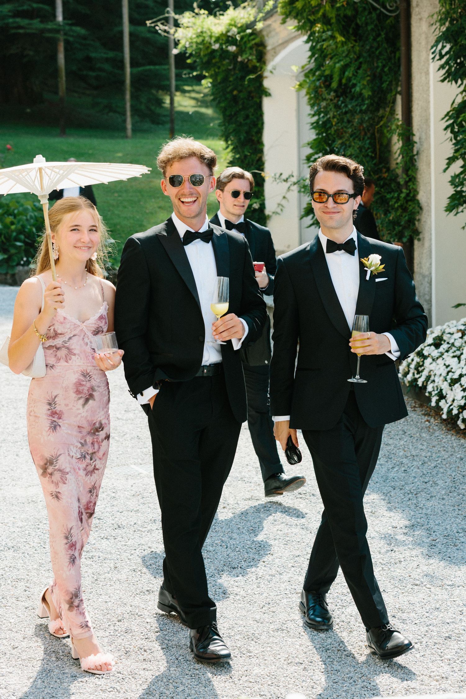 Wedding guests in tuxedos and floral dress walk outdoors holding champagne glasses at an elegant garden celebration.