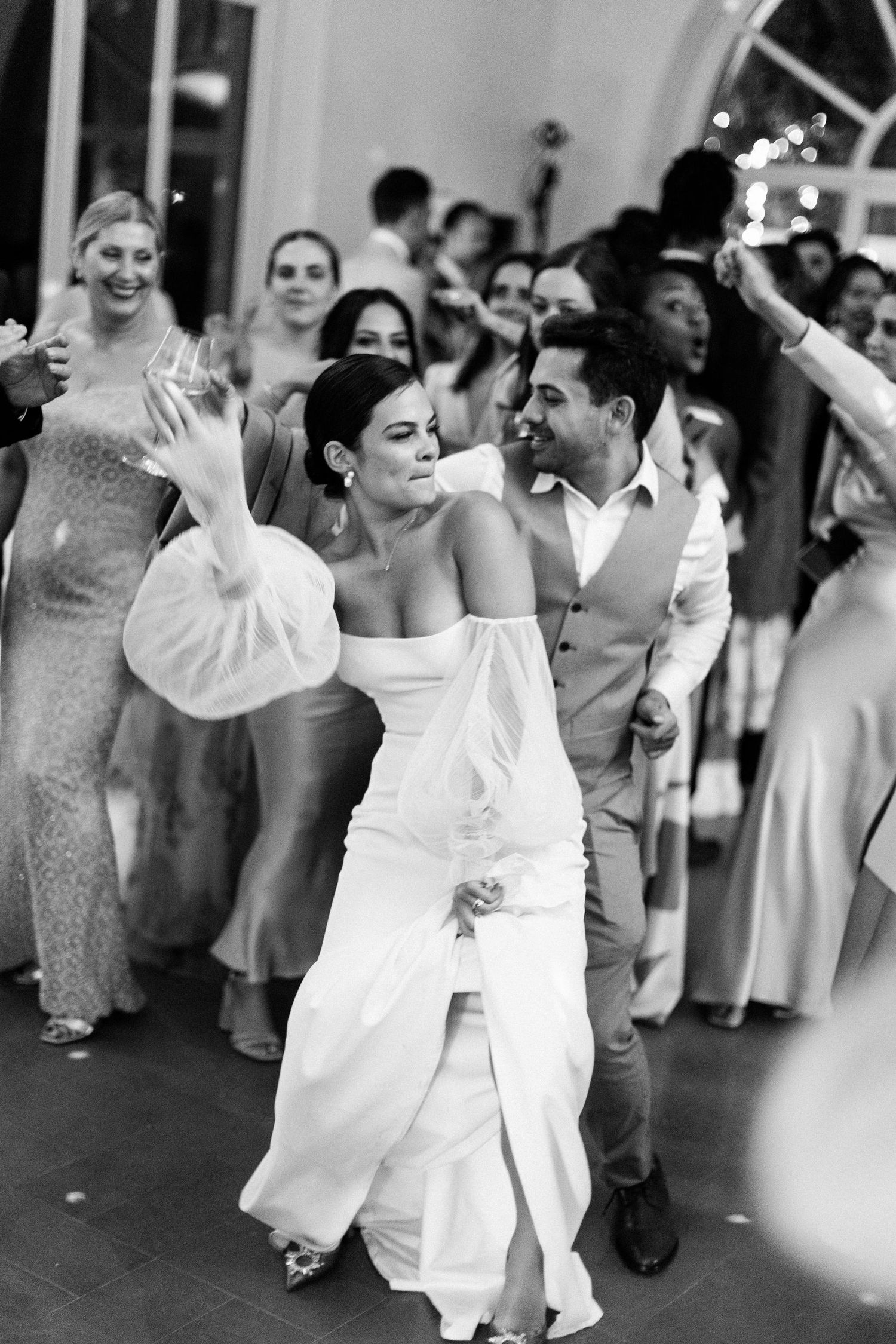 Bride in off-shoulder white gown dances joyfully with guests celebrating at elegant wedding reception.